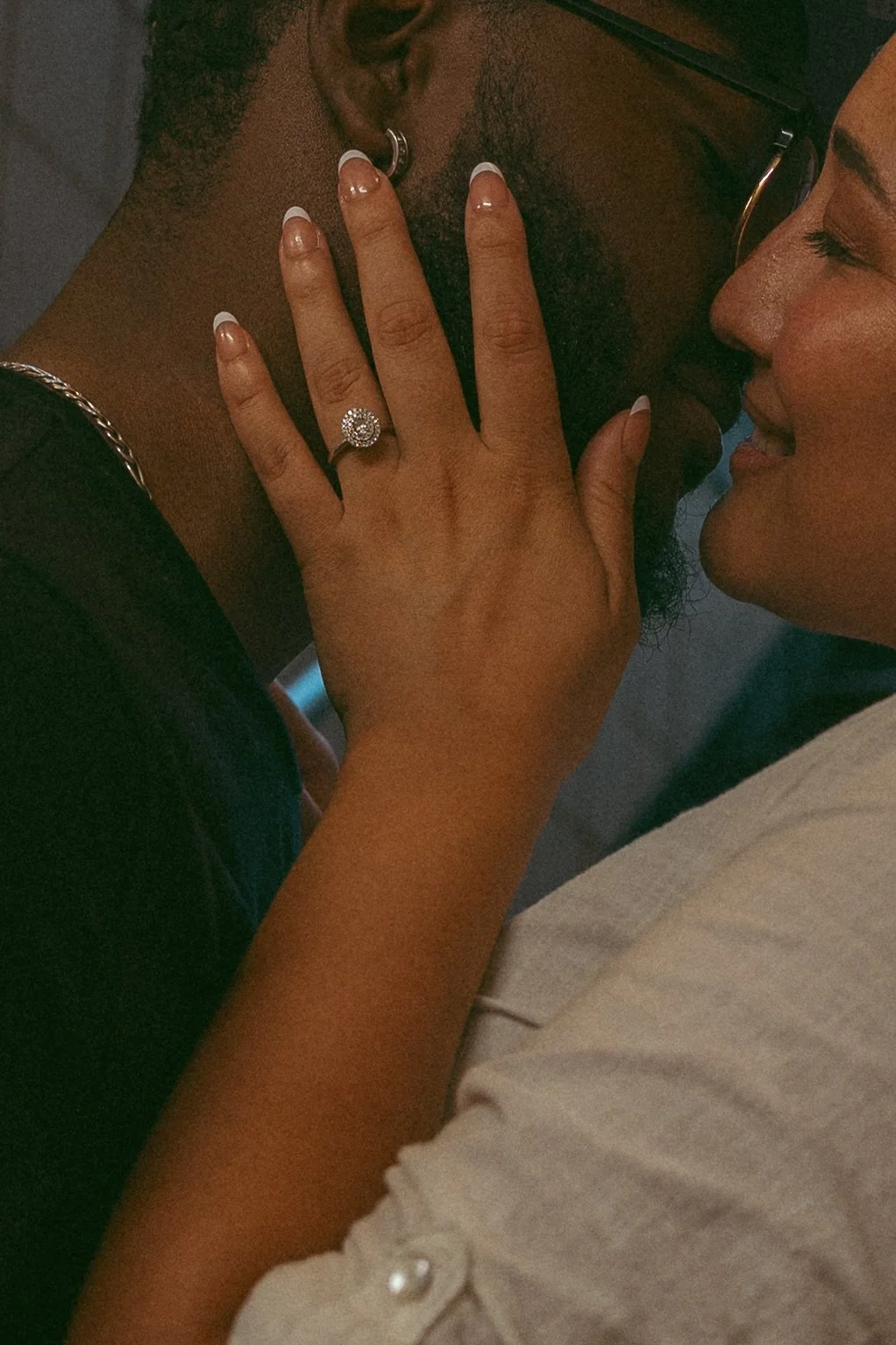 Two women are embracing closely, with one woman’s hand resting on the other’s chest, displaying an engagement ring with a prominent diamond. The woman on the right has her eyes closed and is smiling, while the woman on the left is wearing a dark top with a pearl detail on the sleeve.