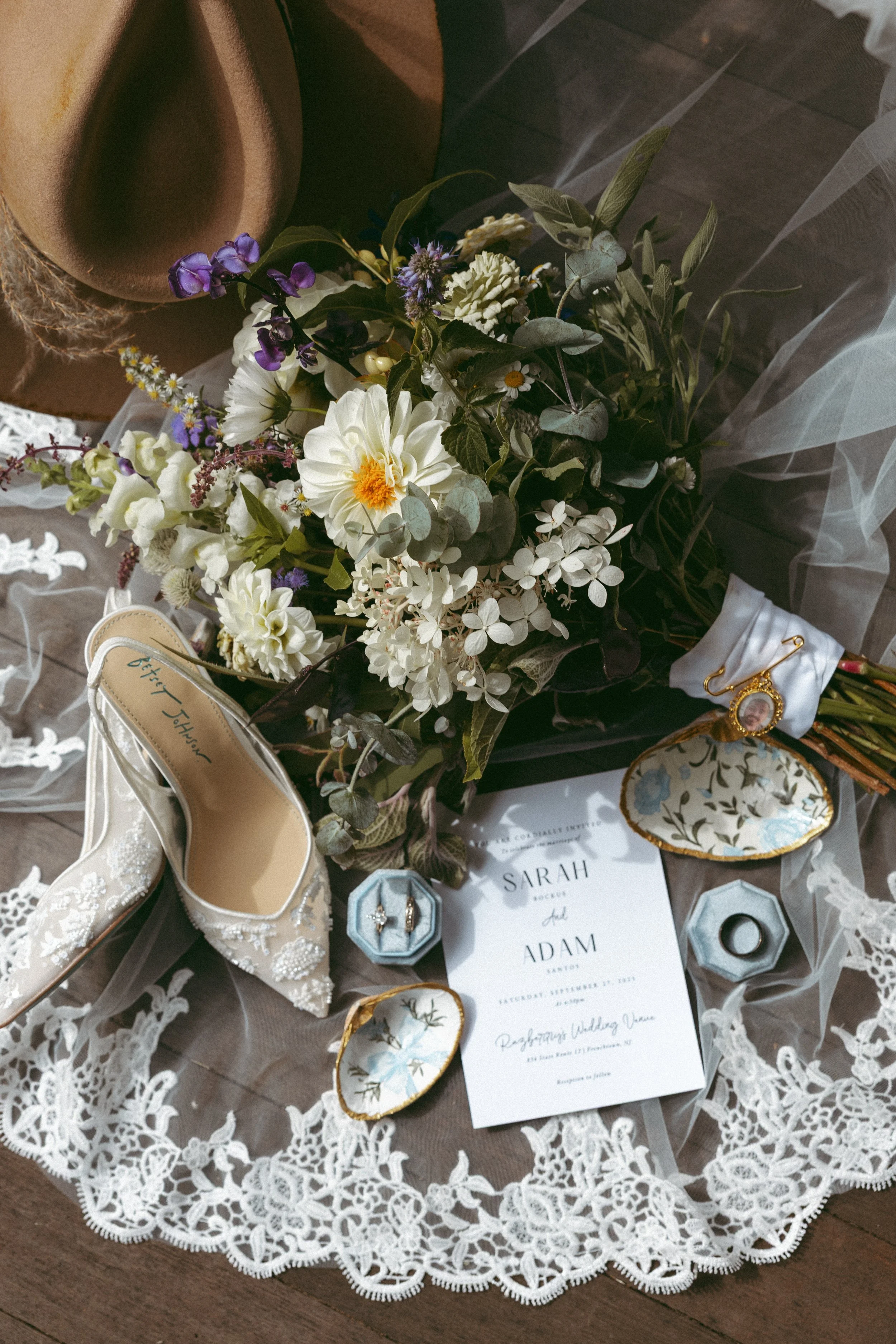 Wedding invitation, floral bouquet, wedding shoes, jewelry, and accessories arranged on lace fabric.