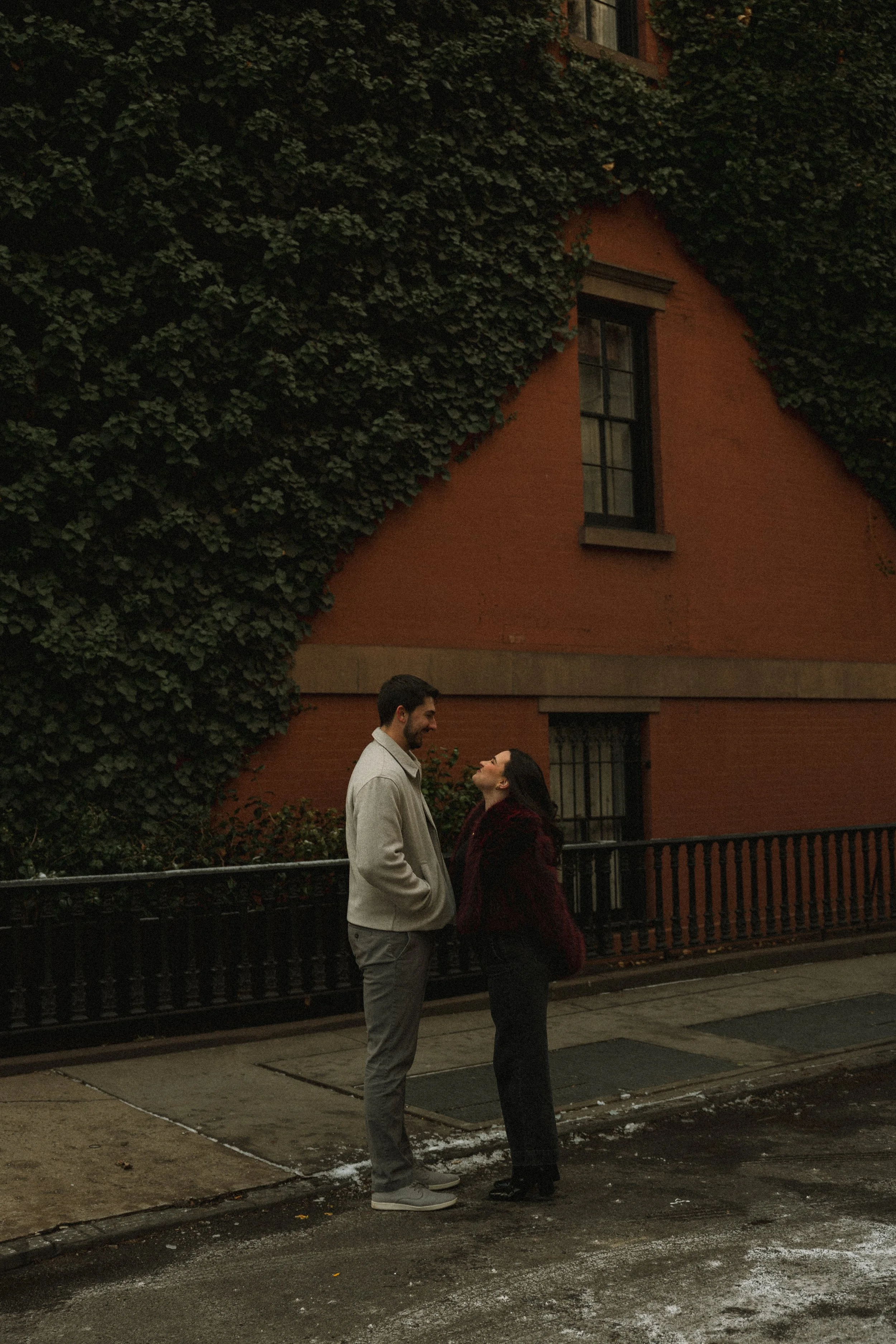 A couple standing on a street at dusk, facing each other and smiling, with a large ivy-covered building in the background