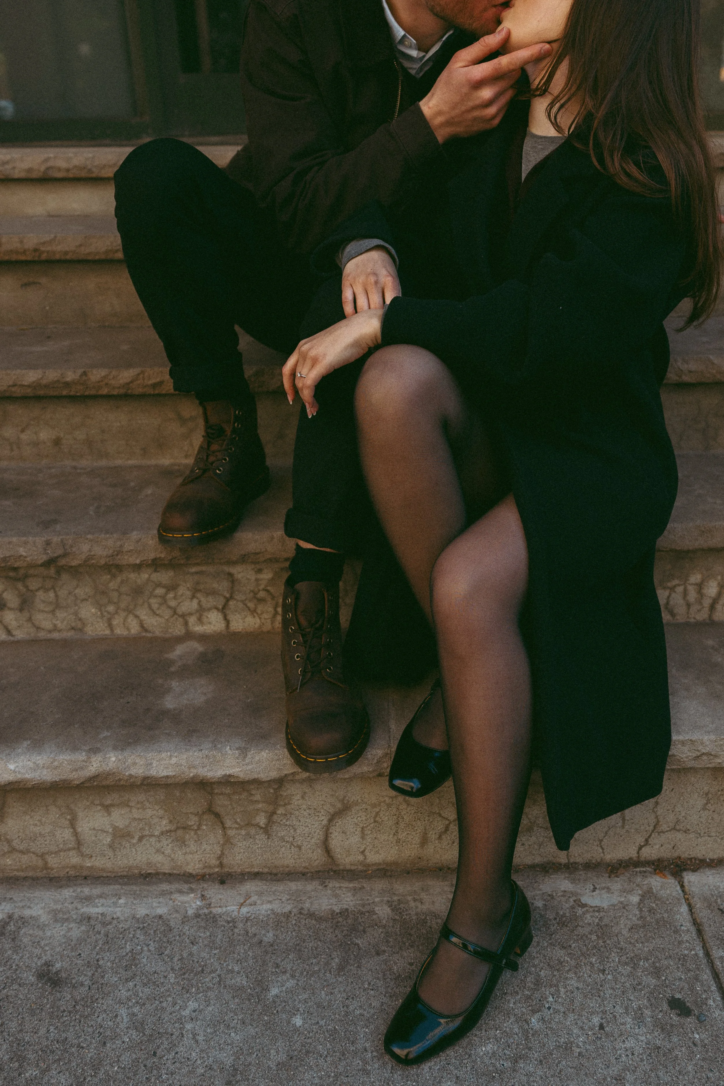 A couple sitting on stone stairs, engaged in a romantic kiss. The woman is dressed in black with sheer tights and open-toe shoes, and the man wears dark clothing with boots.