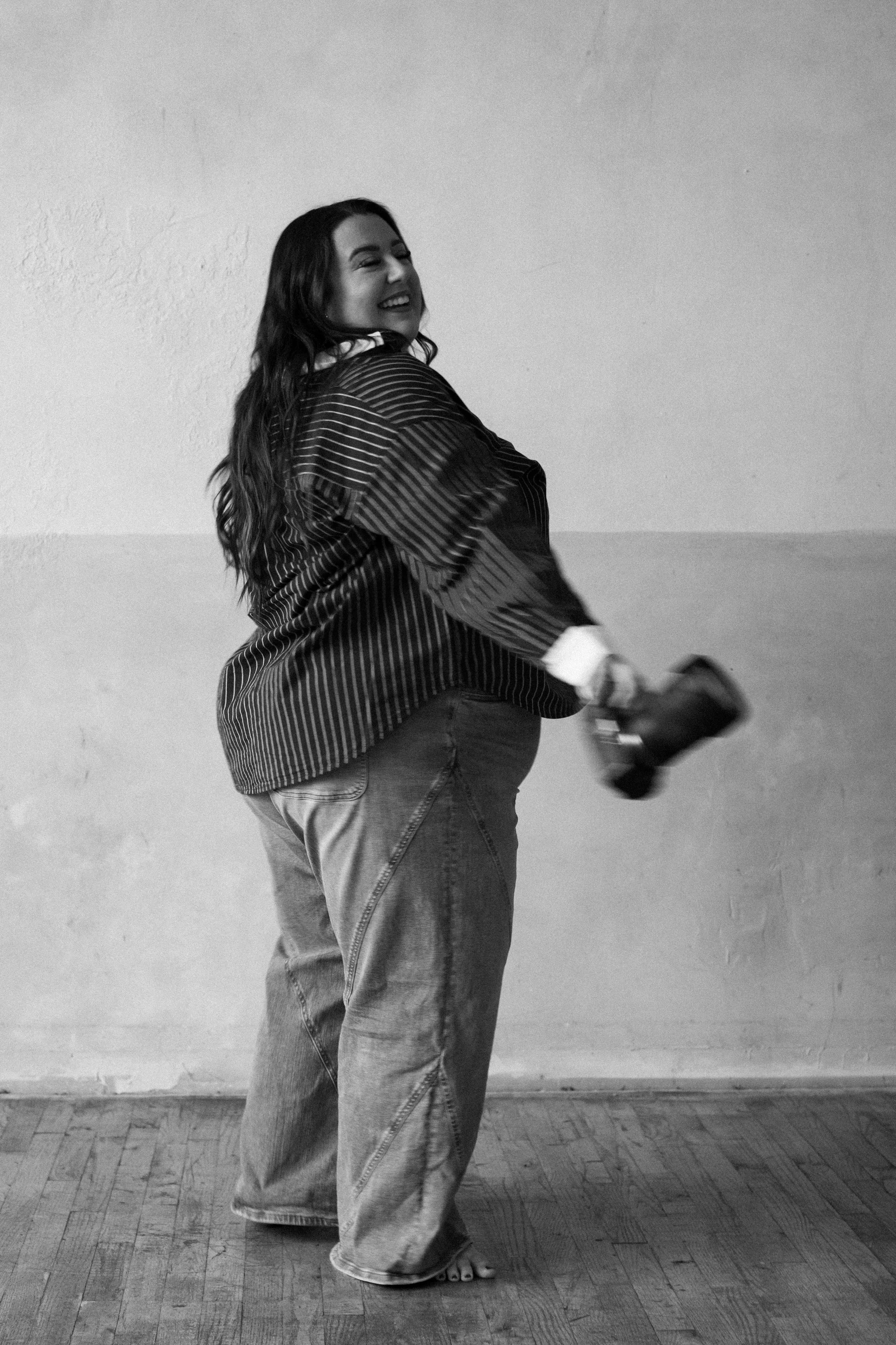A smiling woman with long dark hair, wearing a striped shirt and loose jeans, standing barefoot on a wood floor, holding a camera, against a plain wall.