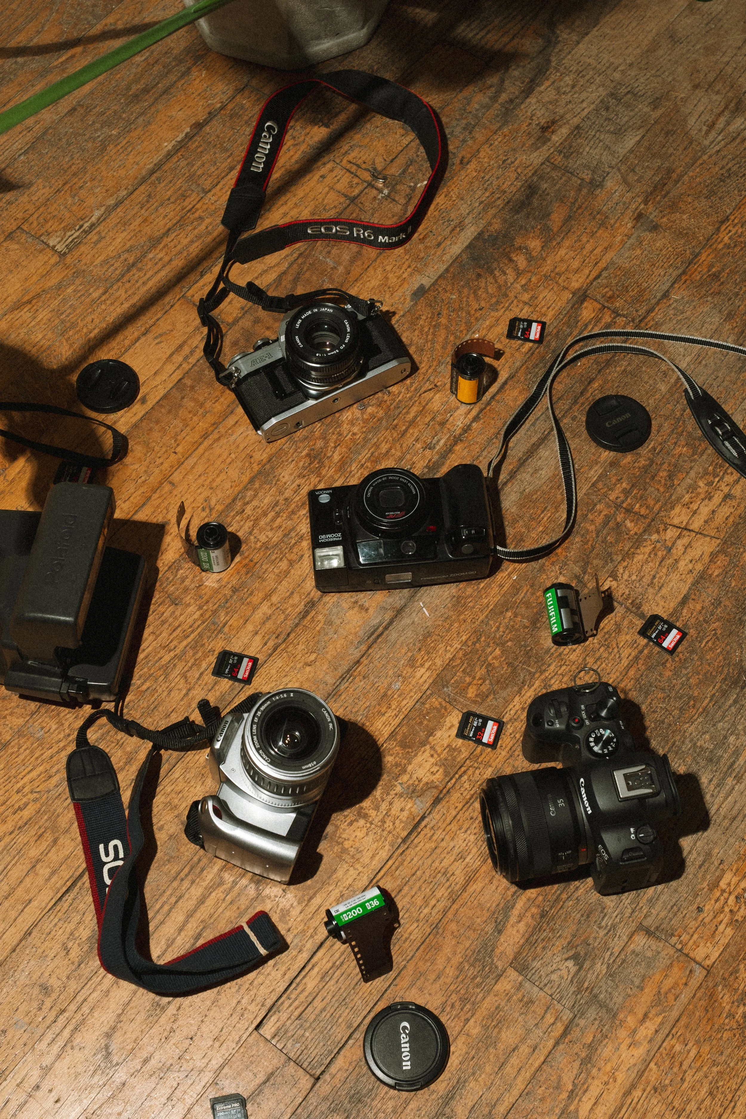 Various cameras, batteries, memory cards, lens caps, and camera accessories scattered on a wooden floor.
