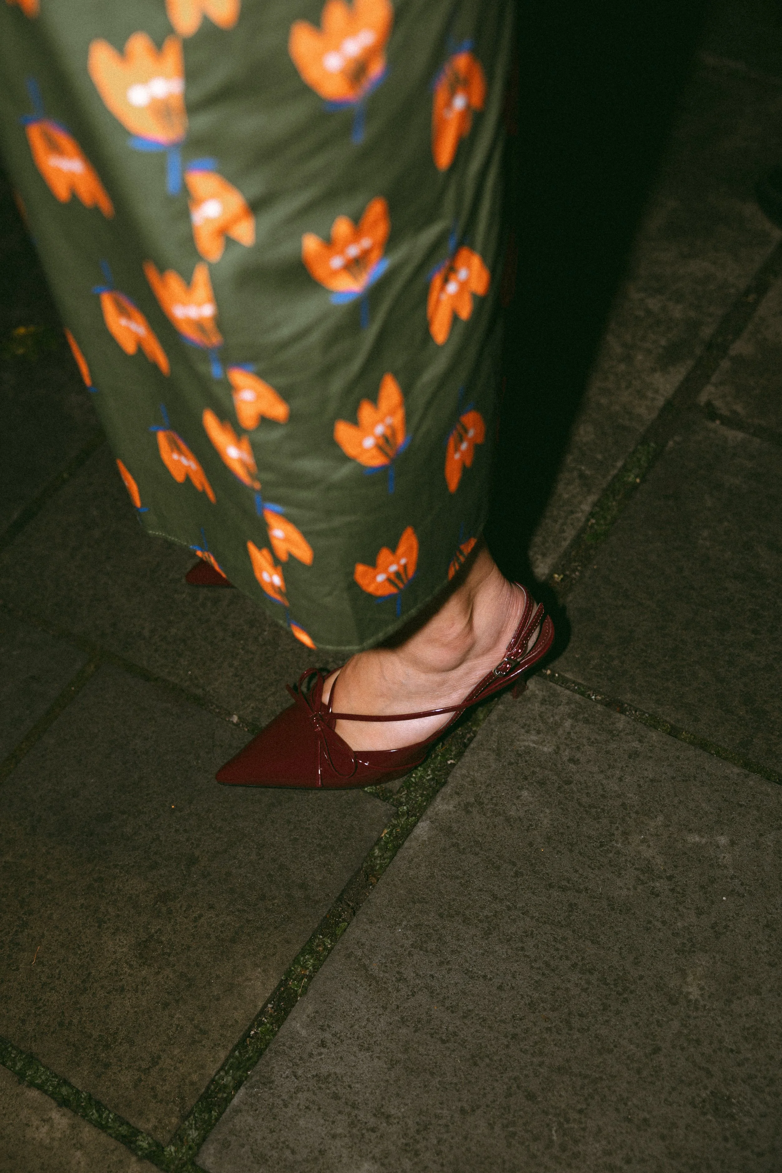 Close-up of a person's foot in a burgundy high heel shoe with a pointed toe and ankle strap, on a dark stone pavement, partially covered by a green skirt with a floral pattern of orange flowers with blue stems.
