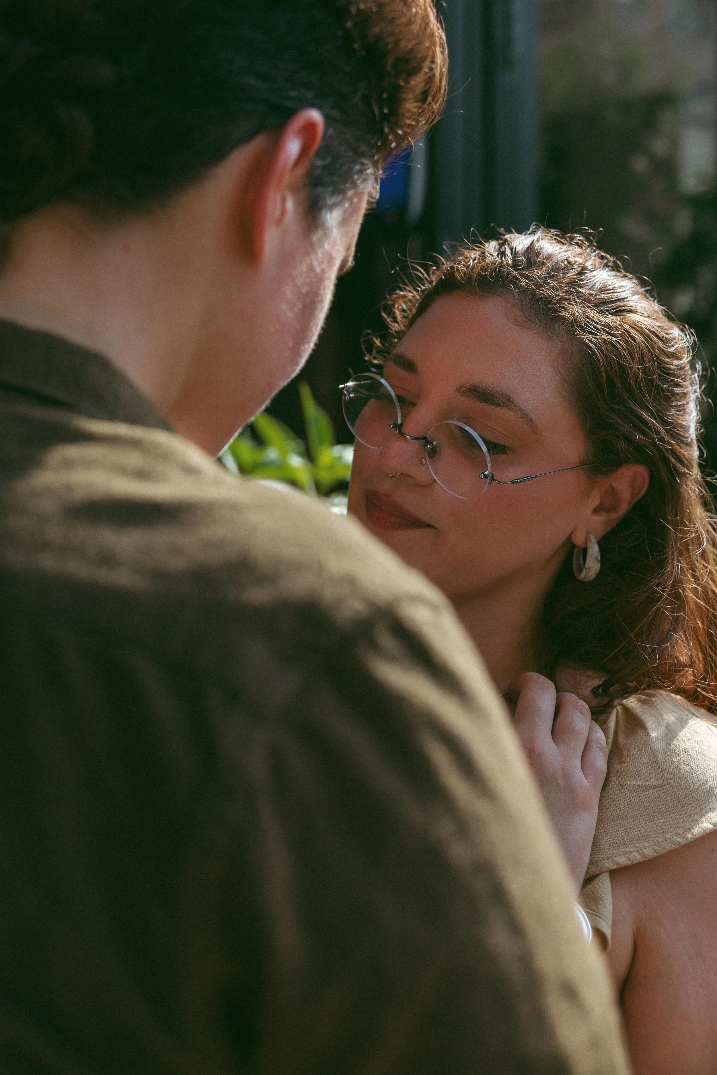 A woman with glasses and piercings being close to a man, with the woman touching her shoulder and looking at the man.