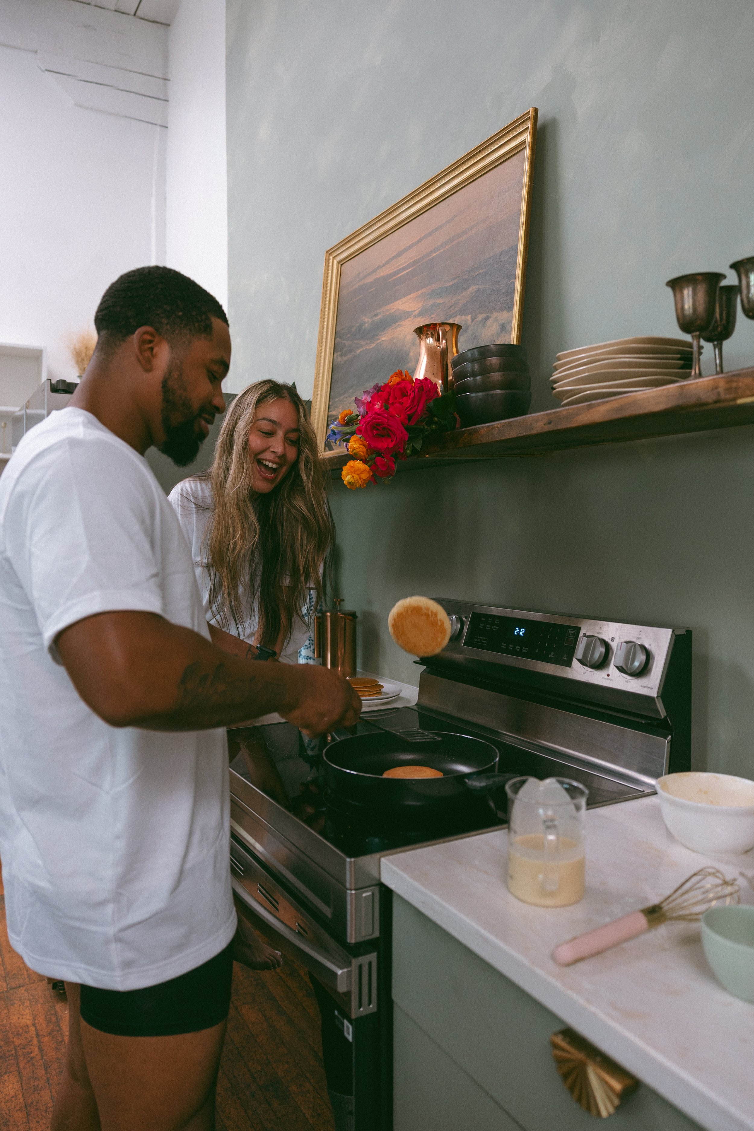 A man and woman cooking breakfast together in a kitchen, with a man flipping a pancake in a pan and a woman smiling nearby.