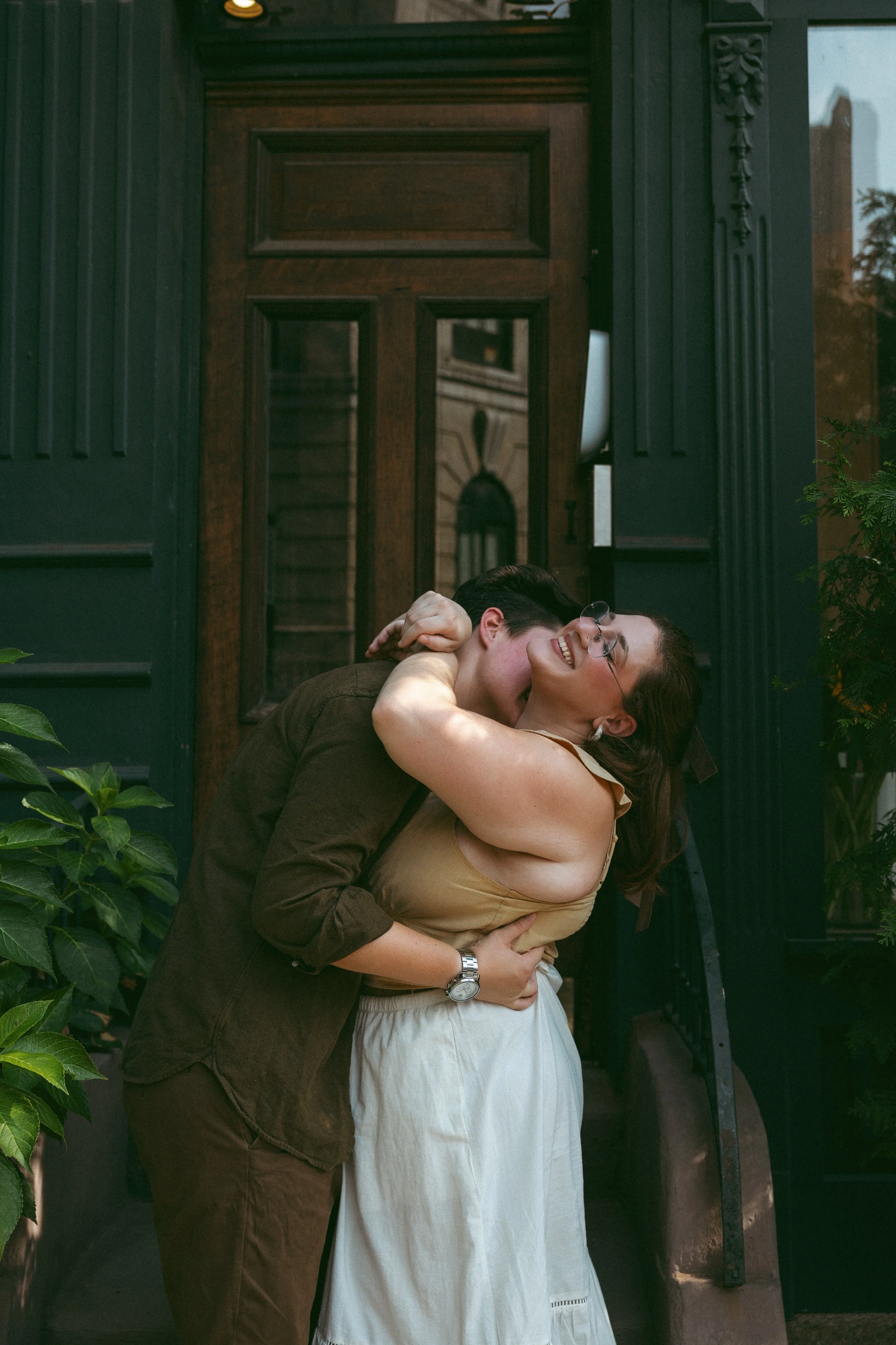 A couple hugging and smiling in front of a house entrance, surrounded by greenery.