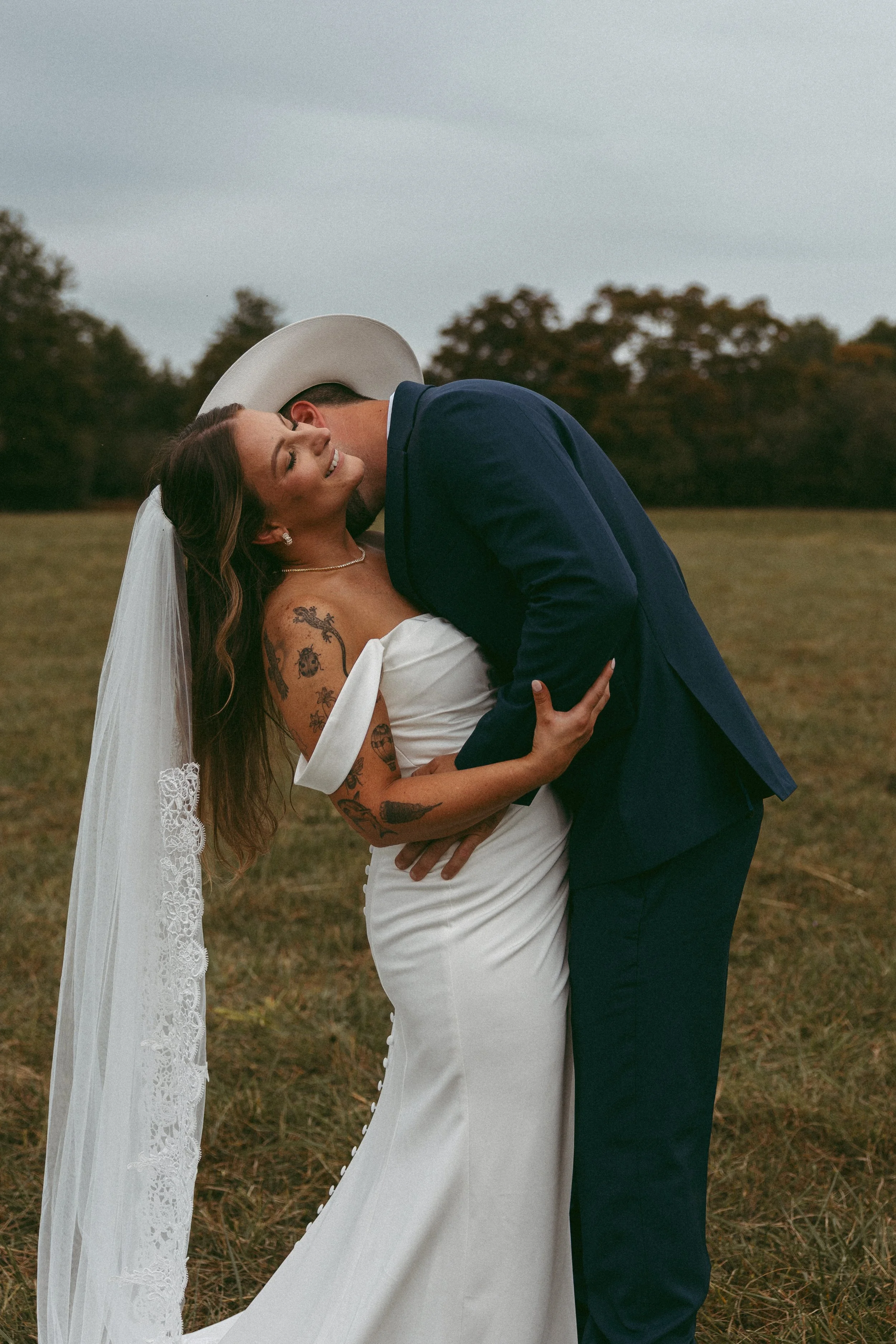 A newlywed couple, a woman in a white wedding dress with tattoos and a veil, and a man in a dark blue suit and cowboy hat, sharing a romantic moment outdoors on a grassy field with trees in the background.