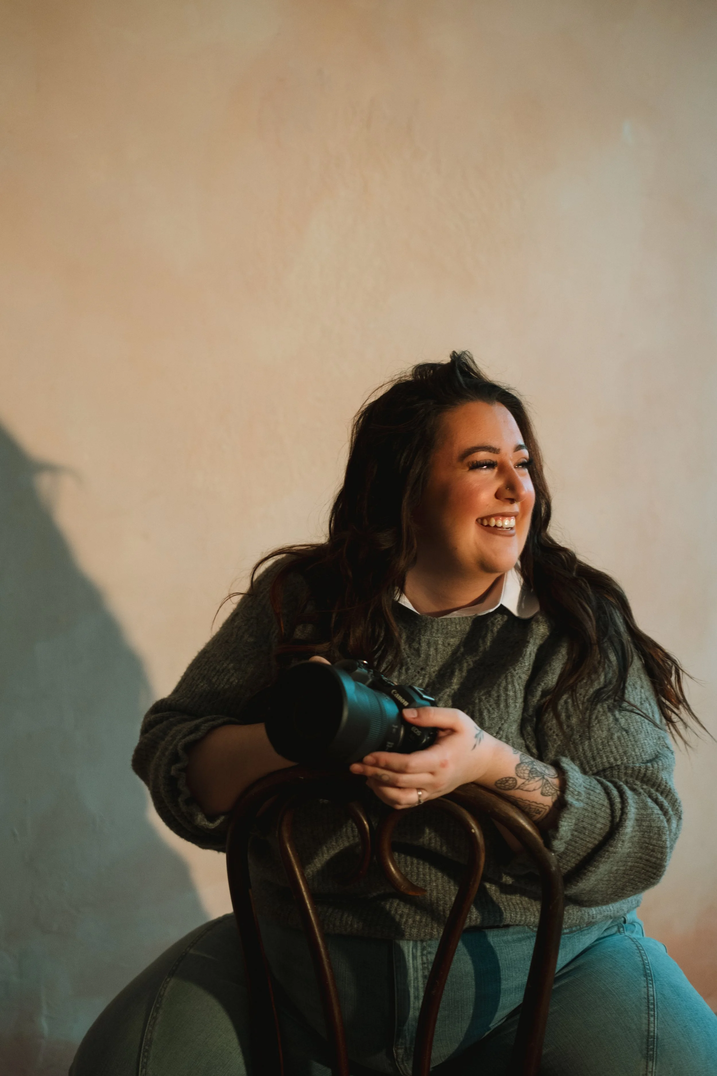 A smiling woman with dark wavy hair, tattoos on her arm, wearing a gray sweater and jeans, sitting on a chair, holding a camera, in front of a plain wall with warm lighting.