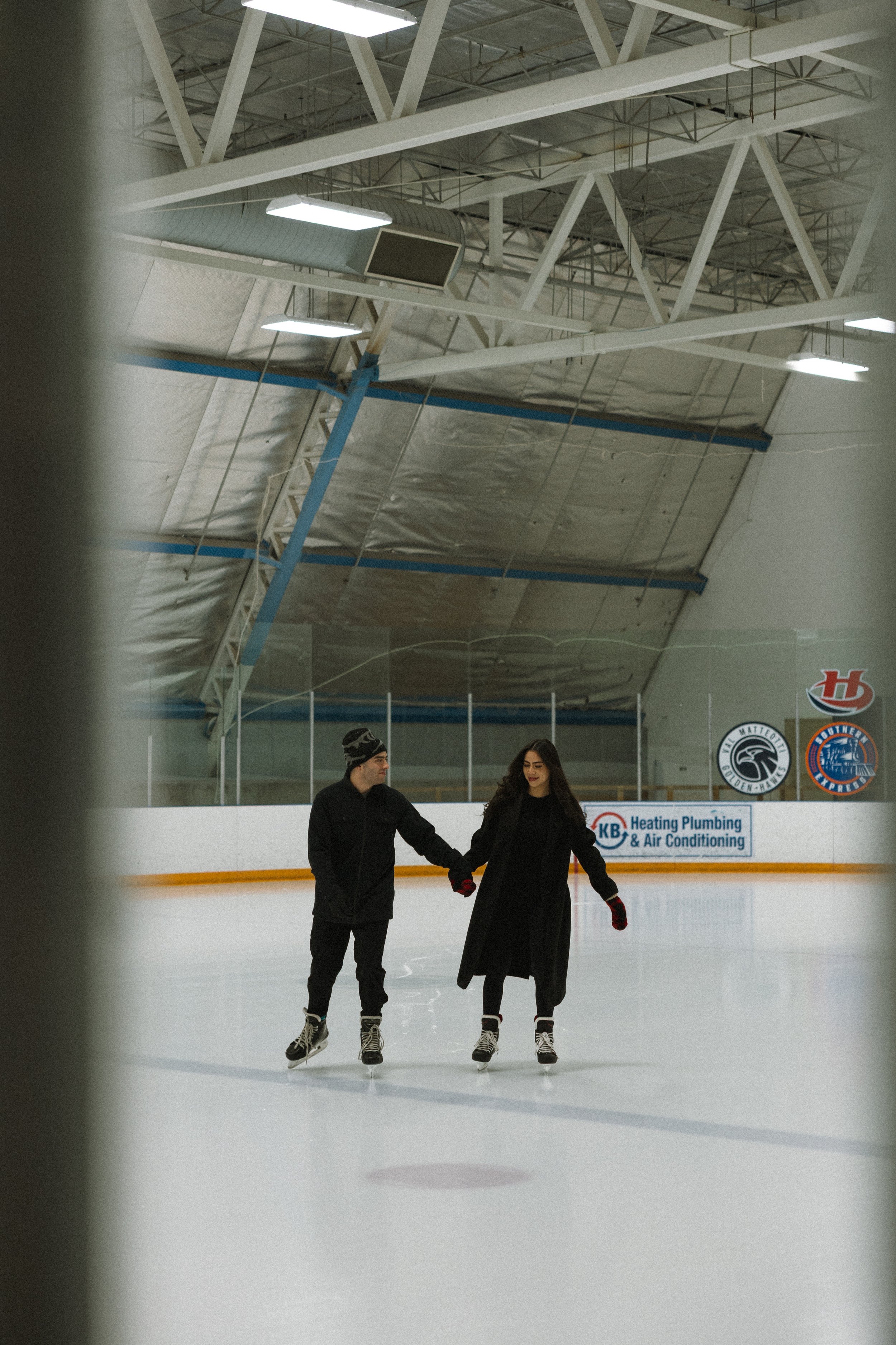 A man and woman ice skating inside an indoor ice rink, holding hands and skating together.