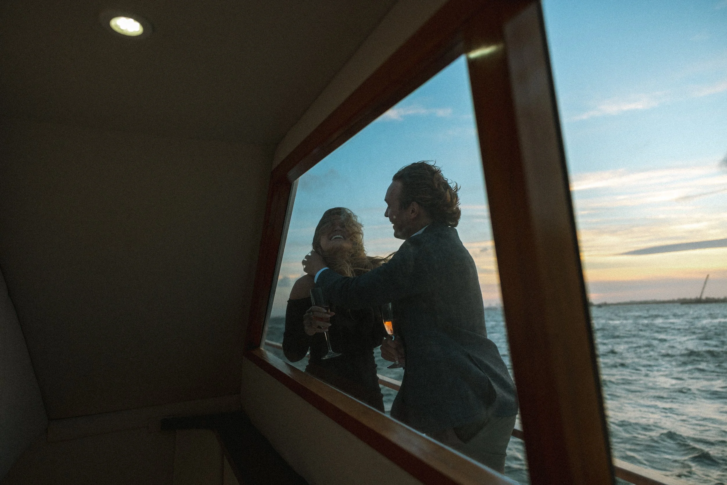 A man and woman in formal attire sharing a laughing moment on a boat, viewed through a window with a scenic water and sky background during sunset.