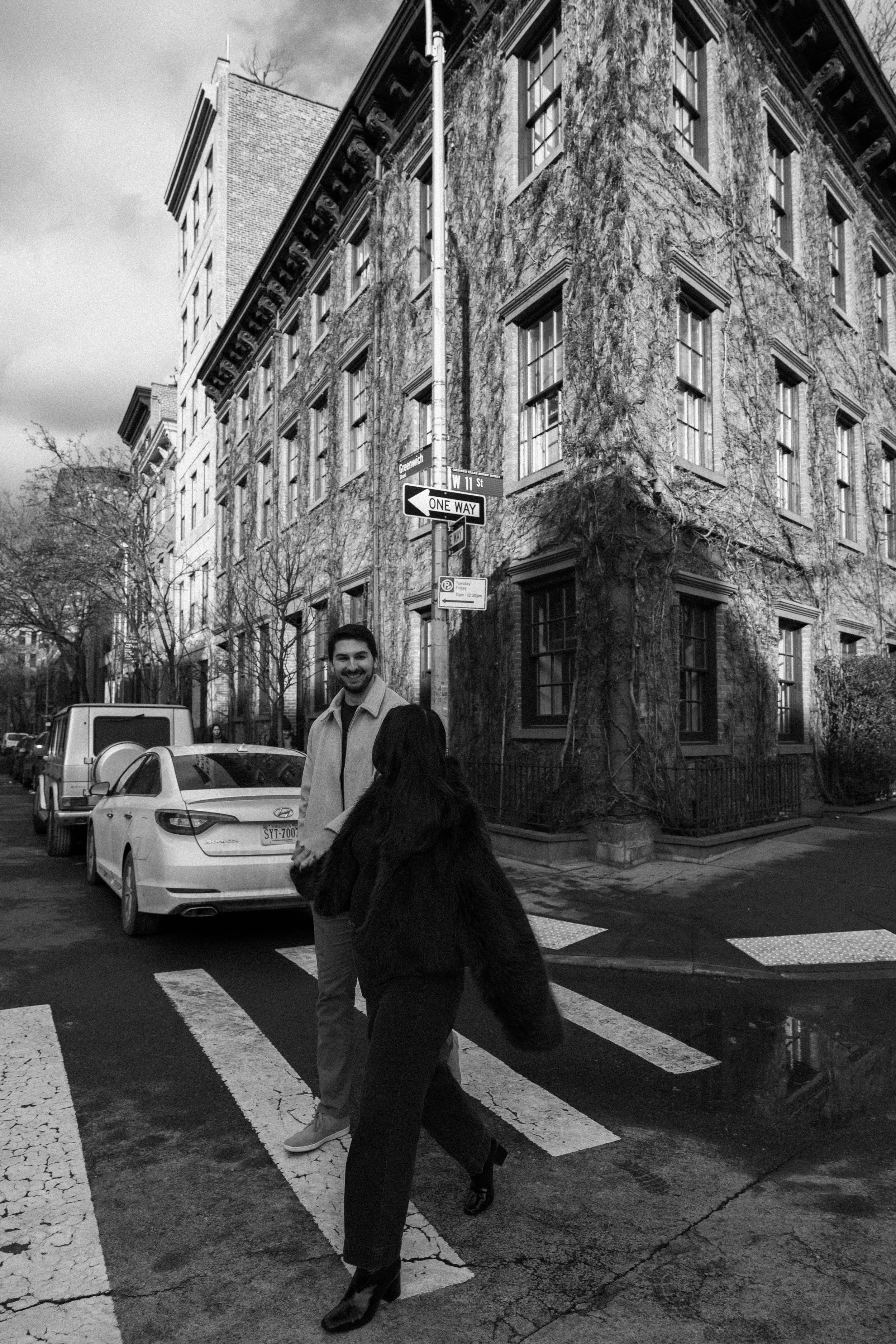 Two people crossing a city street at a pedestrian crosswalk, with parked cars and an old brick building covered in ivy in the background, in black and white.