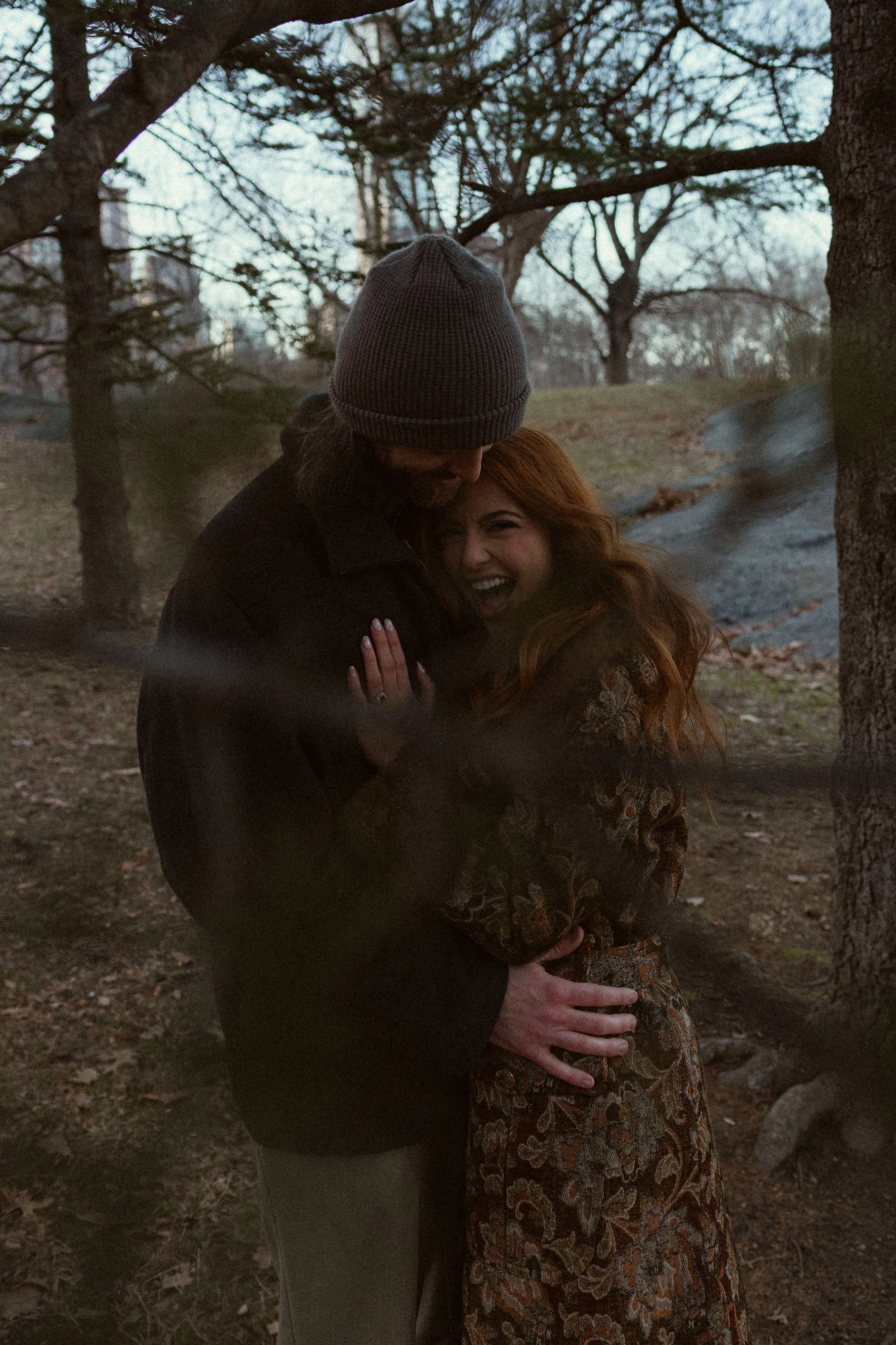A man and a woman embrace outdoors among trees, smiling and appearing happy.