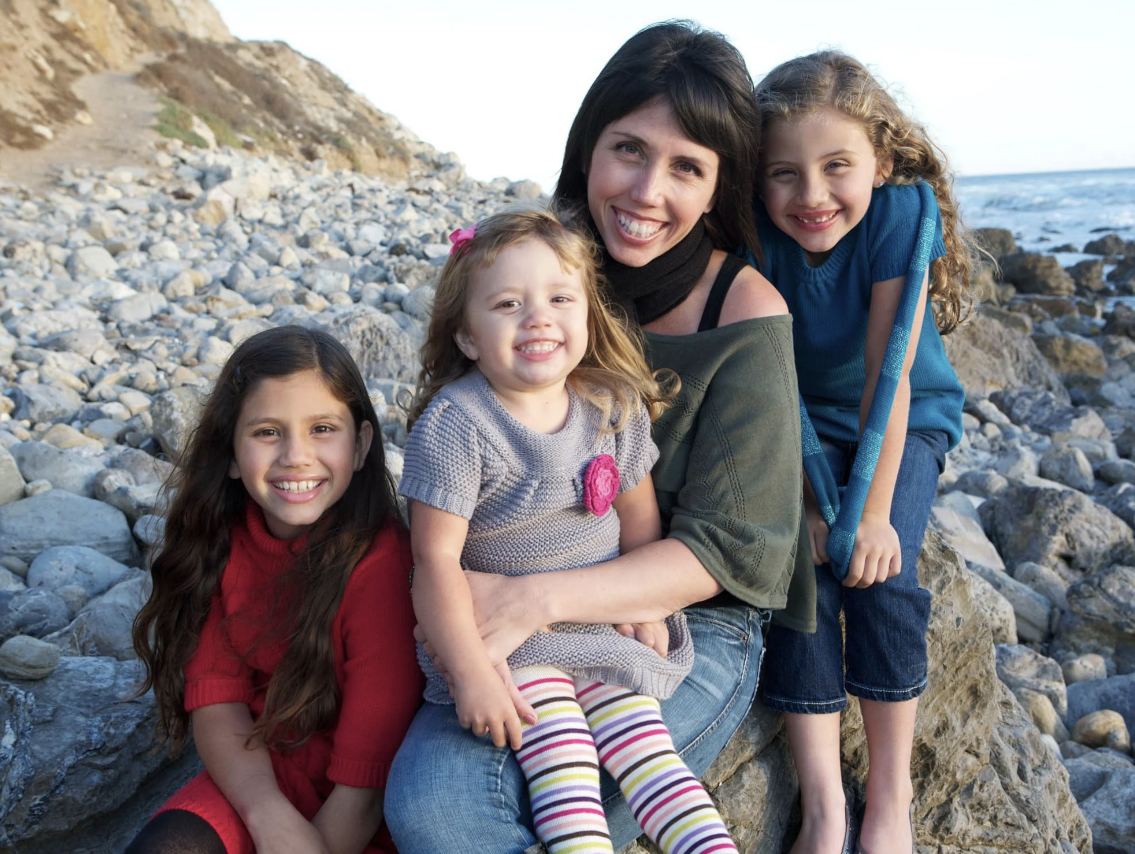 A woman and four young girls sitting on rocks at a beach, smiling, with cliffs and the ocean in the background.
