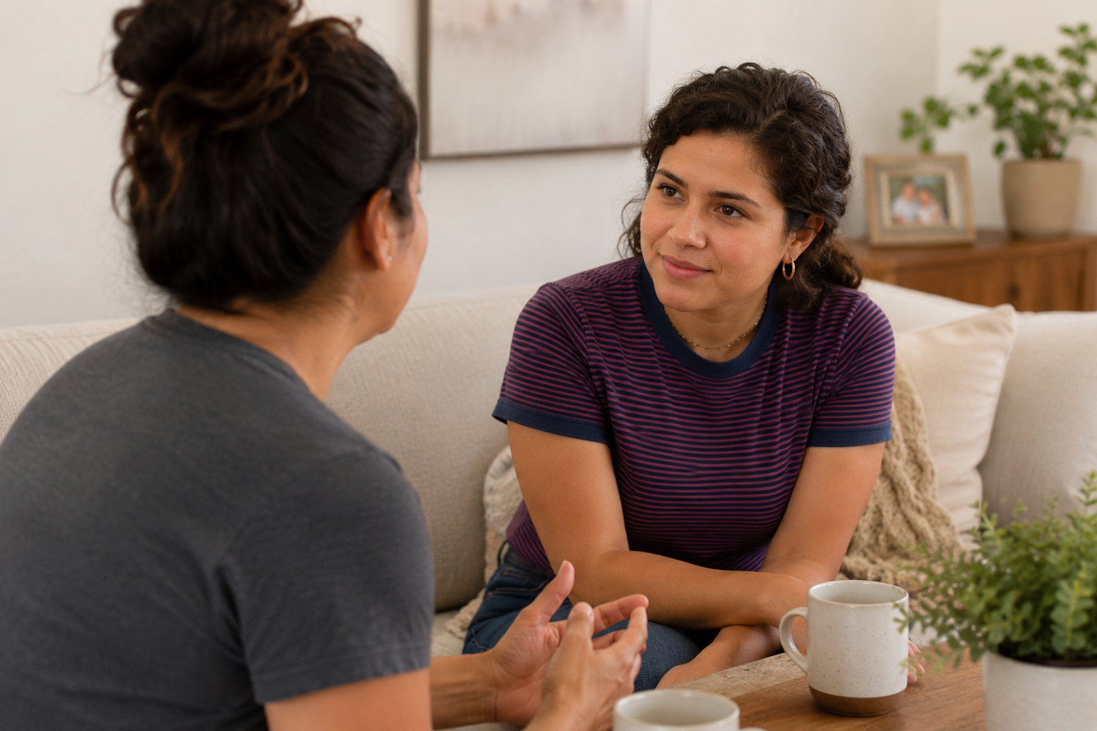 Two women sit on a beige sofa having a conversation, with coffee mugs on a wooden table in front of them. One woman has dark hair styled in a bun, and the other has short, curly hair. There are framed photos and a plant in the background.