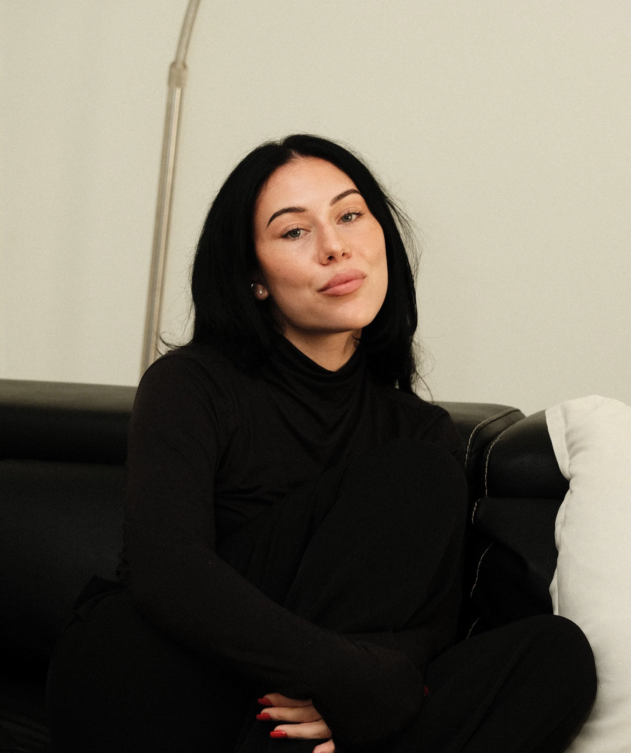 A woman with black hair, wearing a black turtleneck, sitting on a black sofa with white pillows in a living room.