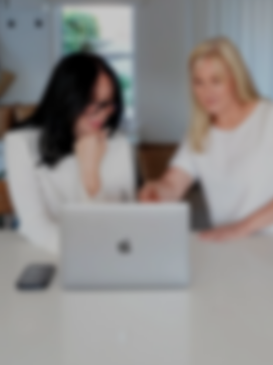 Two women sitting at a table with a laptop and a smartphone, engaging in a discussion in a bright room.