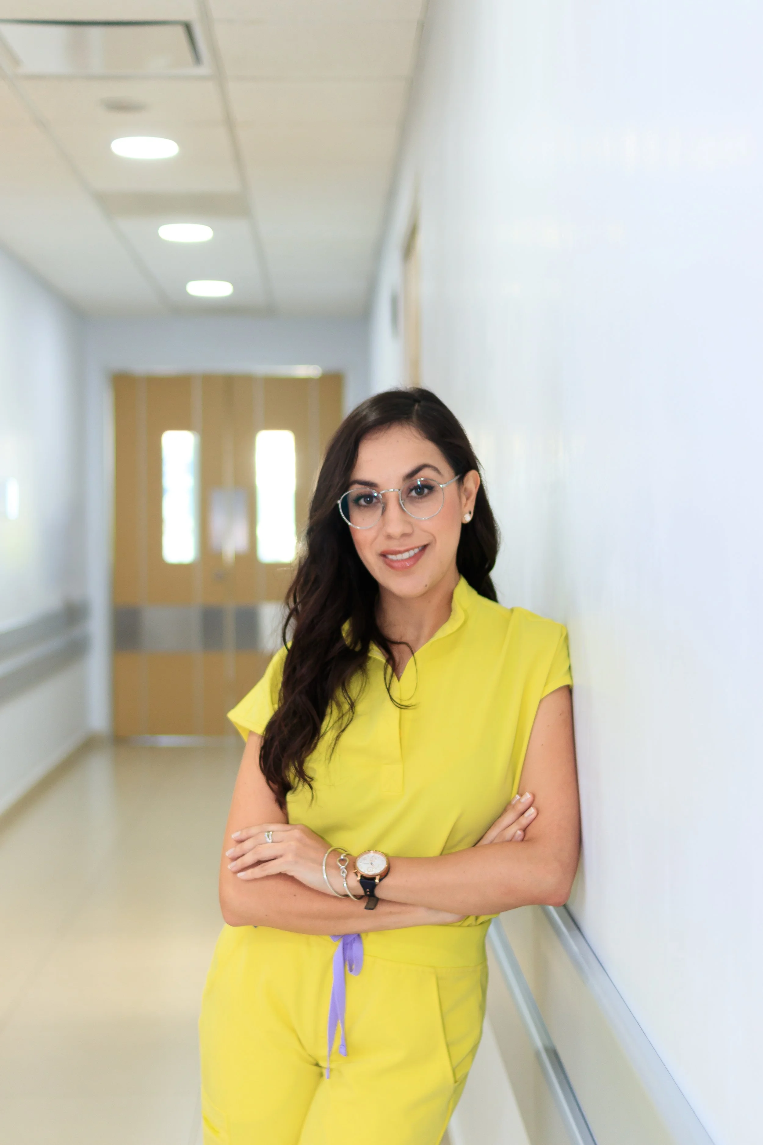 Mujer joven con gafas, de cabello largo y oscuro, vestida con ropa amarilla, sonriendo y apoyada en la pared en un pasillo con puertas al fondo.