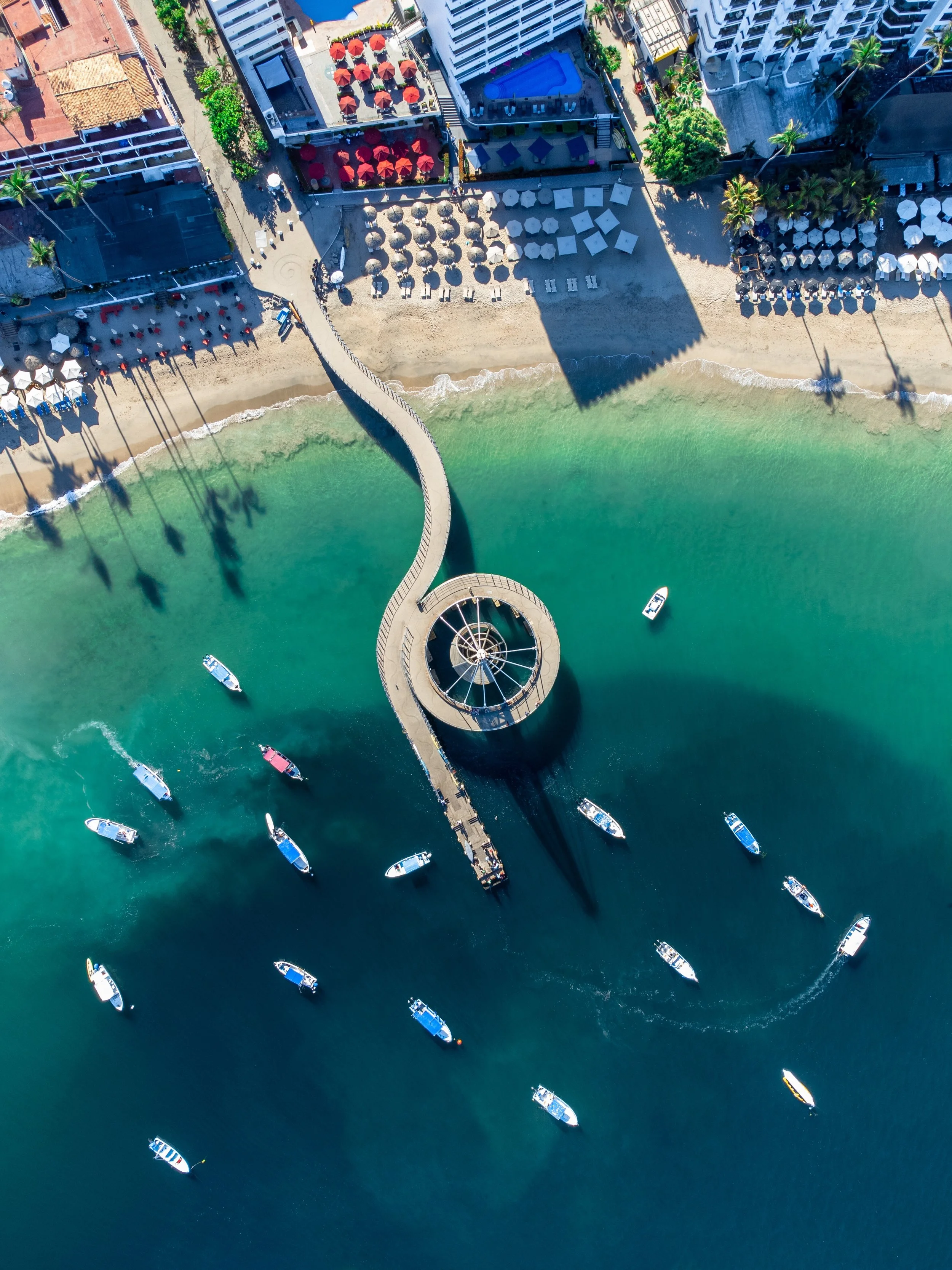 Vista aérea de una playa con un muelle curvado que llega al mar, varios barcos en el agua y sombrillas en la arena, con edificios y áreas de descanso cercanas.