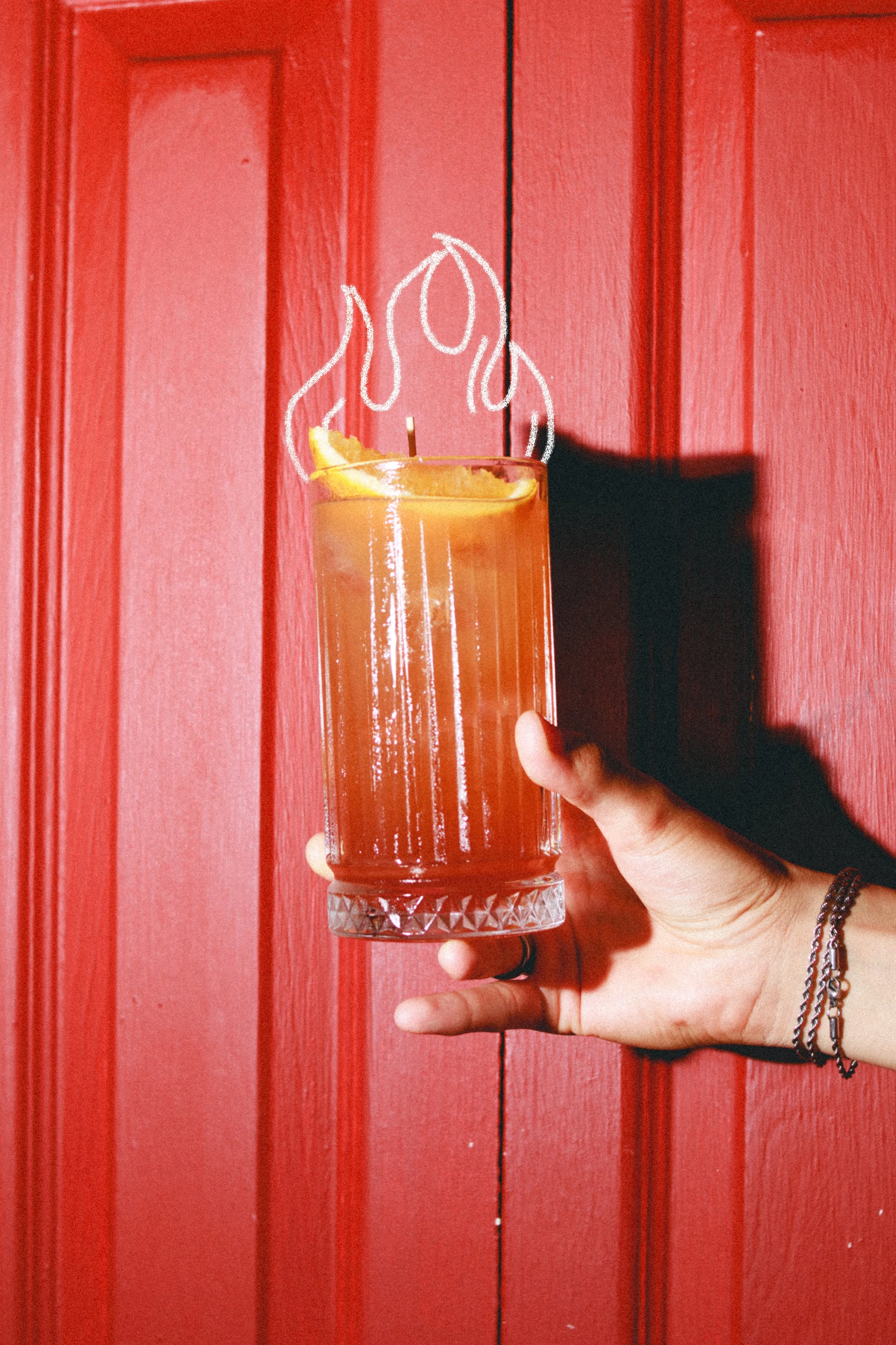 Mano sosteniendo un vaso con una bebida amarilla y una flor de limón, con dibujo de llamas blancas sobre la bebida, frente a una puerta roja.