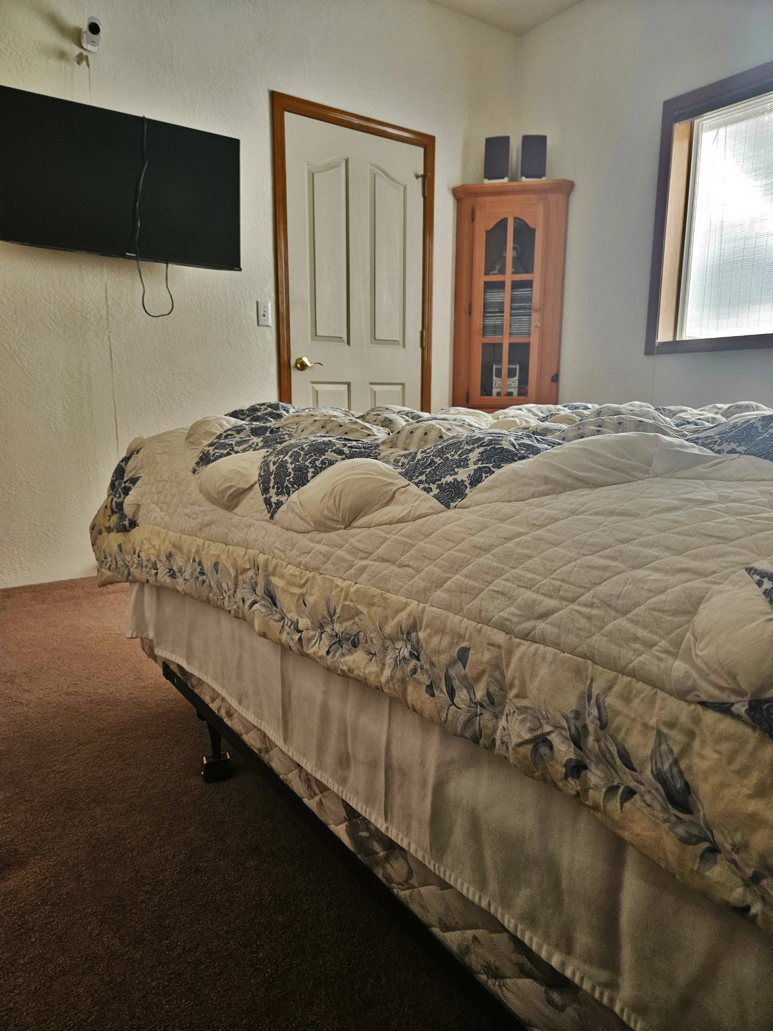 A bedroom with a bed covered in a floral quilt, a wall-mounted TV with a hanging cord, a wooden cabinet with shelves, a window with blinds, and a closed door.