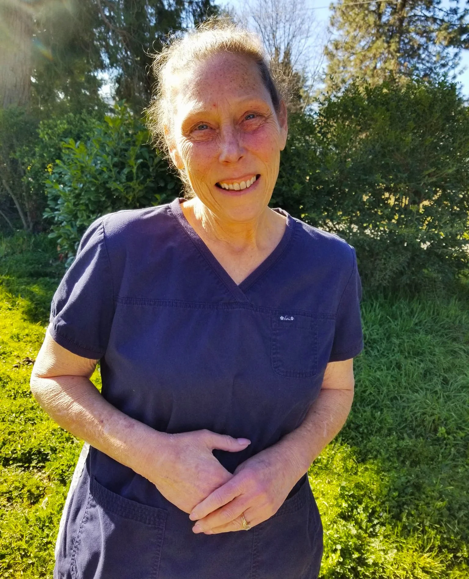 Smiling woman outdoors on a sunny day, wearing navy scrubs, standing in front of green bushes and trees.