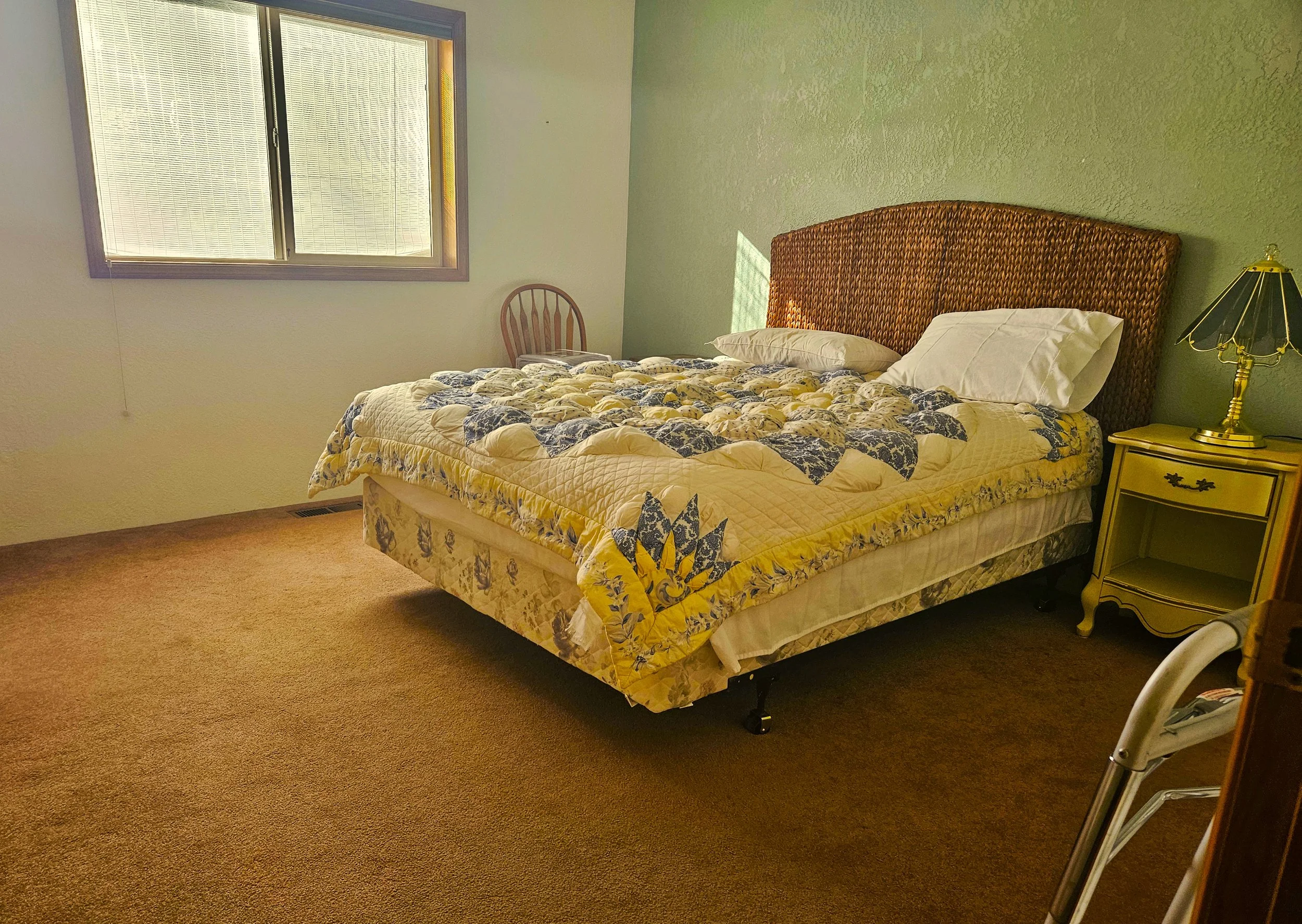 A bedroom with a window, a bed with a floral quilt, white pillows, a wicker headboard, a yellow nightstand with a lamp, a wooden chair, and a bare floor with a vent.