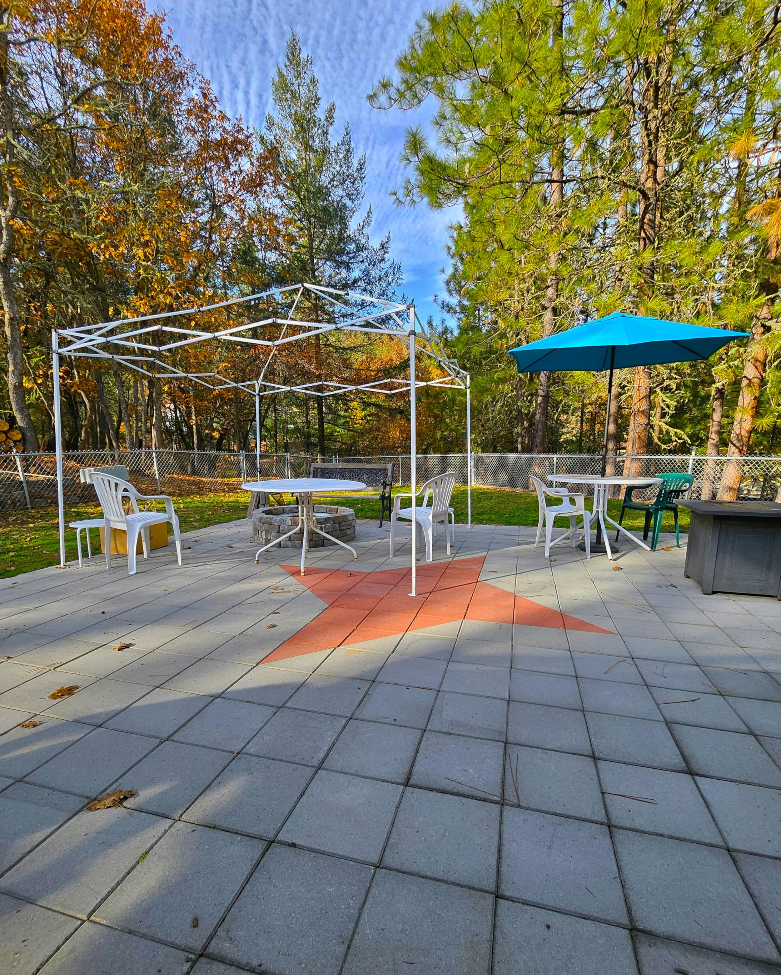 An outdoor patio with a large star design on the tiled floor, surrounded by trees with fall foliage. There are several white plastic chairs, tables, a blue umbrella, a metal fire pit, and a canopy frame without a cover.