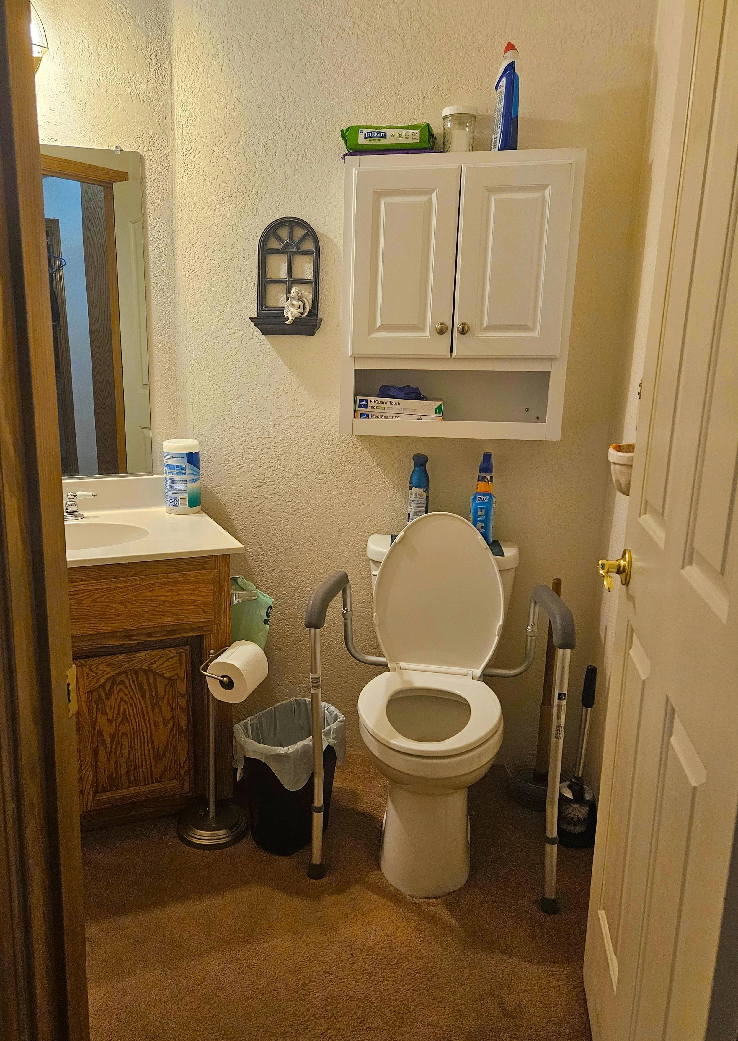 Bathroom with a toilet, a cabinet, and a sink. The toilet has grab bars on each side. Cleaning supplies and personal items are on top of the toilet and cabinet. A mirror is above the sink. A small decorative wall shelf with a figurine is on the wall.