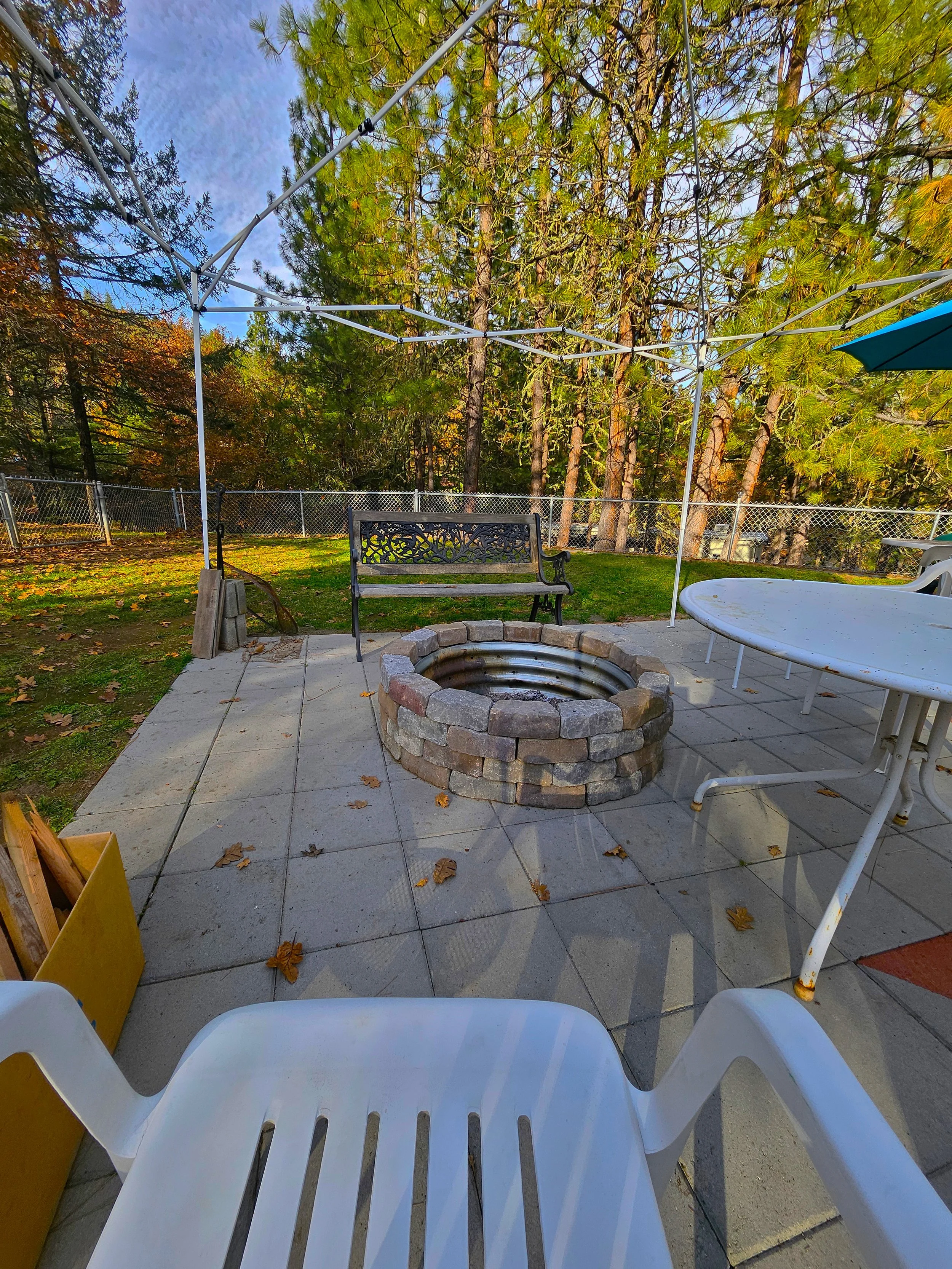 Backyard patio with a fire pit, white plastic chairs, a bench, and a table under a canopy frame, surrounded by trees and a fence.