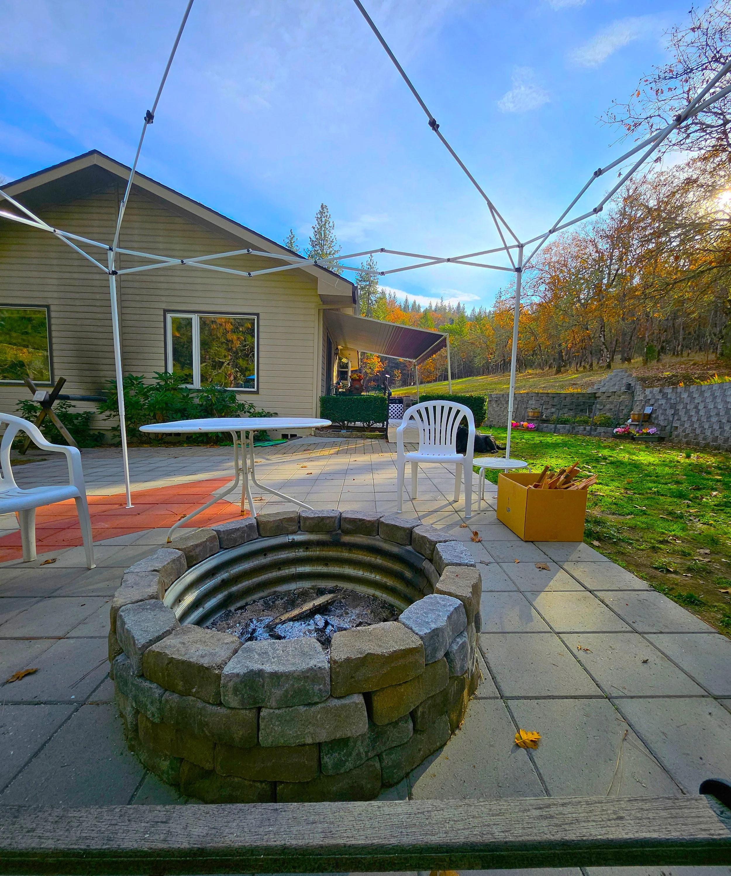 A backyard patio with a fire pit surrounded by plastic chairs, a table, and various outdoor furniture under a canopy frame, with a house and trees in the background.