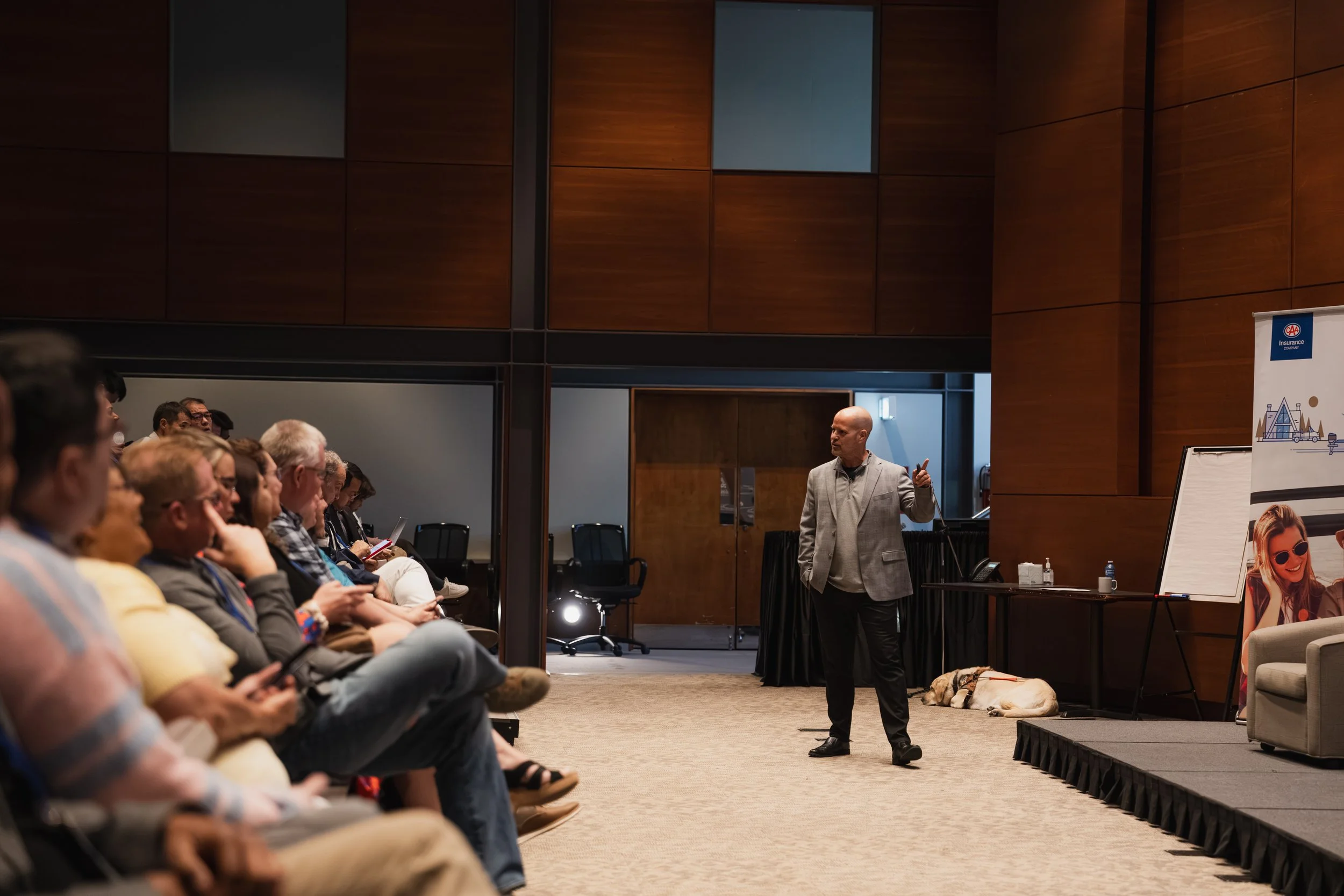 Mark Bentz giving a presentation to an audience seated in a conference room with wood-paneled walls as an engaged audience looks on. His guide dog, Kyle lies on the floor near the stage.
