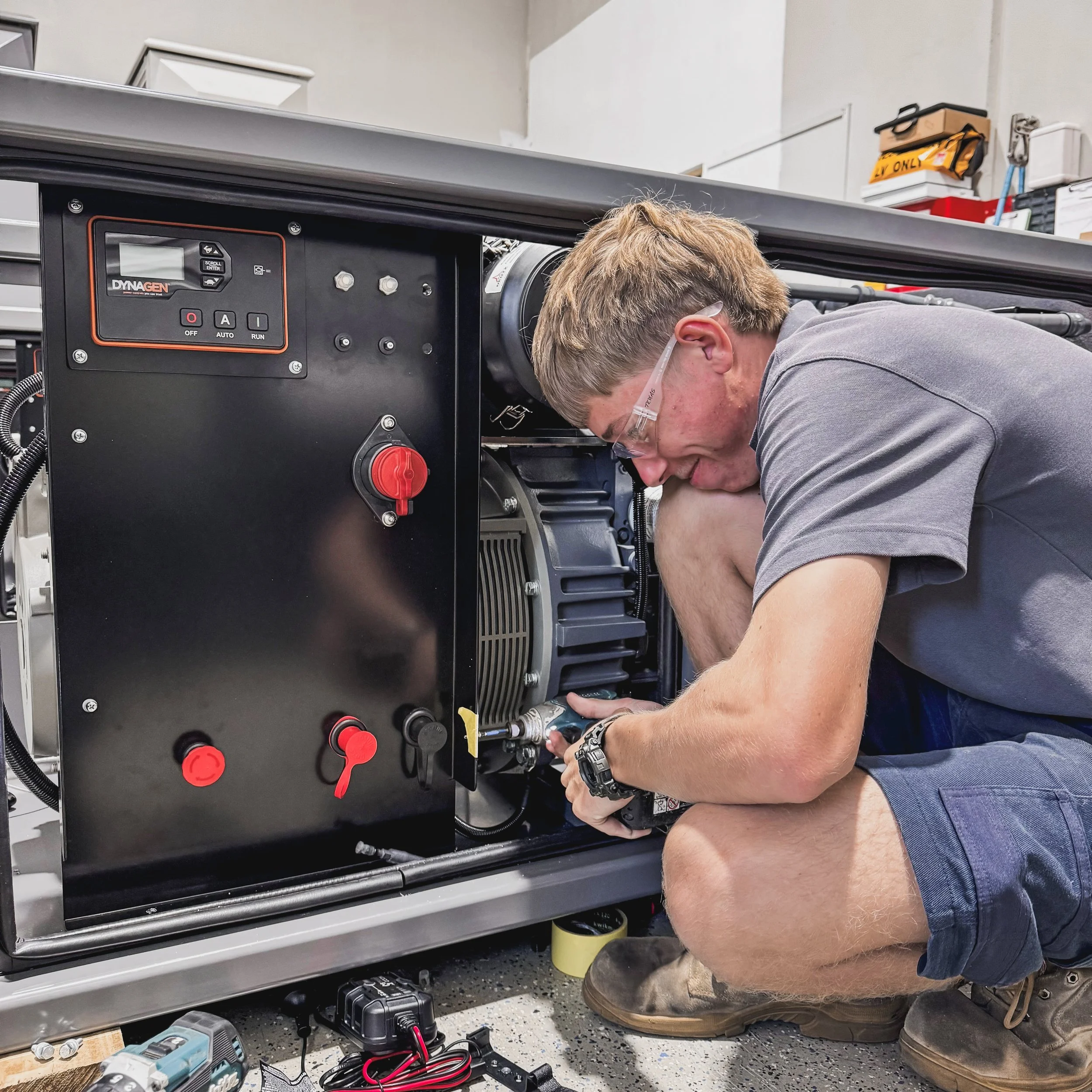 A young man working on a yellow industrial generator or engine, wearing a jacket and shorts, with safety glasses nearby, in a workshop environment.
