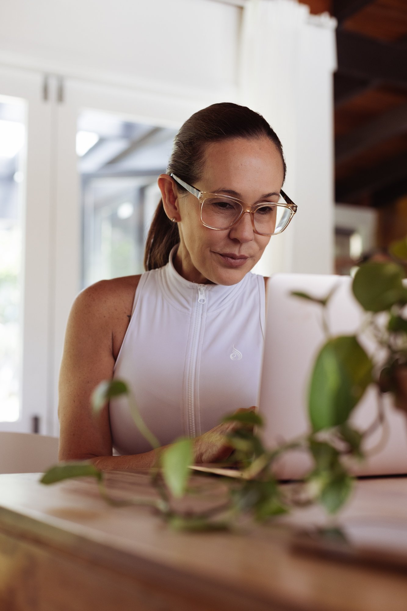 Woman wearing glasses and a white sleeveless zip-up top looking at a laptop, with a plant on the table in front of her, indoors with white walls and a window in the background.