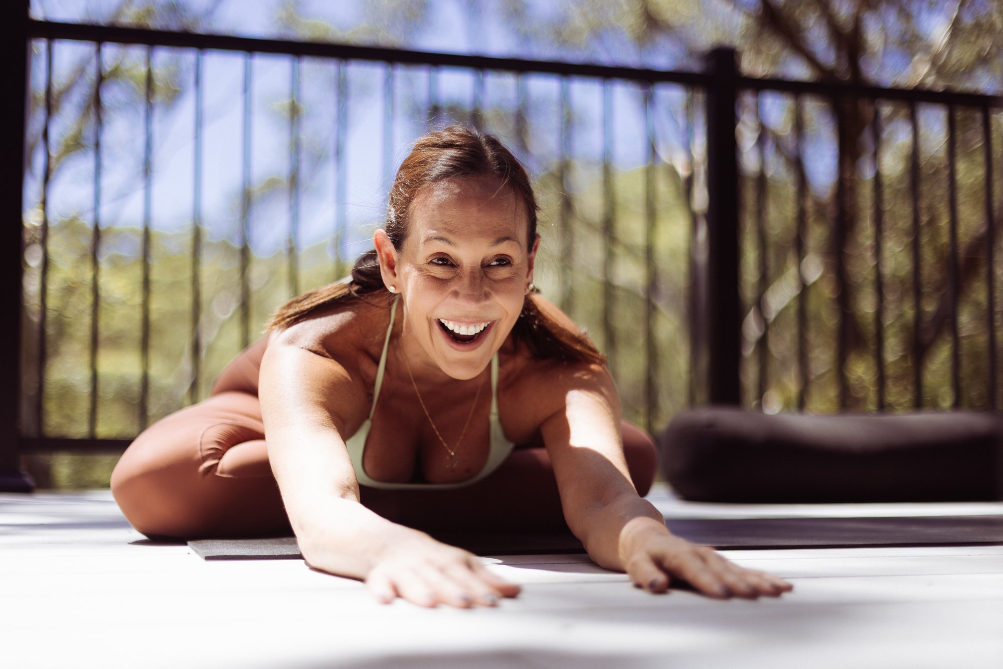 Woman practicing yoga outdoors on a sunny day, smiling and stretching on a yoga mat.