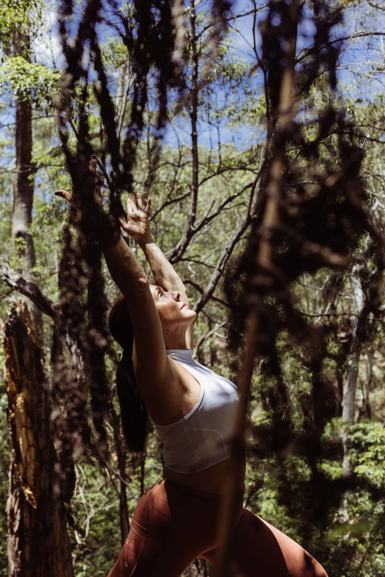 A woman practicing yoga outdoors among trees, stretching her arms upward with a peaceful expression.