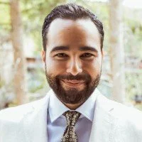 A man with dark hair and a beard smiling, wearing a light-colored suit and tie, outdoors with trees in the background.