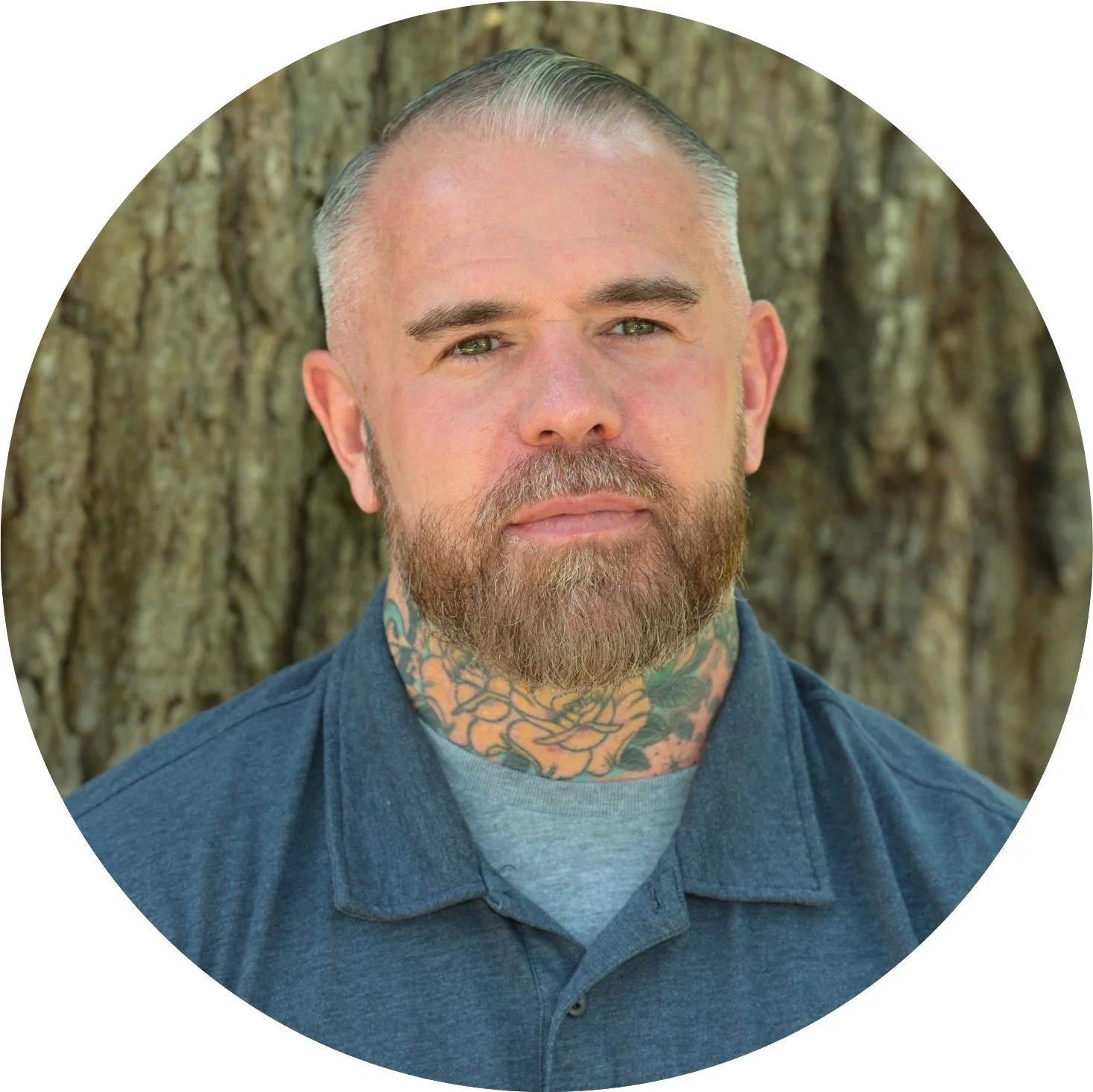 A man with a shaved head, beard, and tattoos on his neck, wearing a blue collared shirt, standing outdoors in front of a tree trunk.