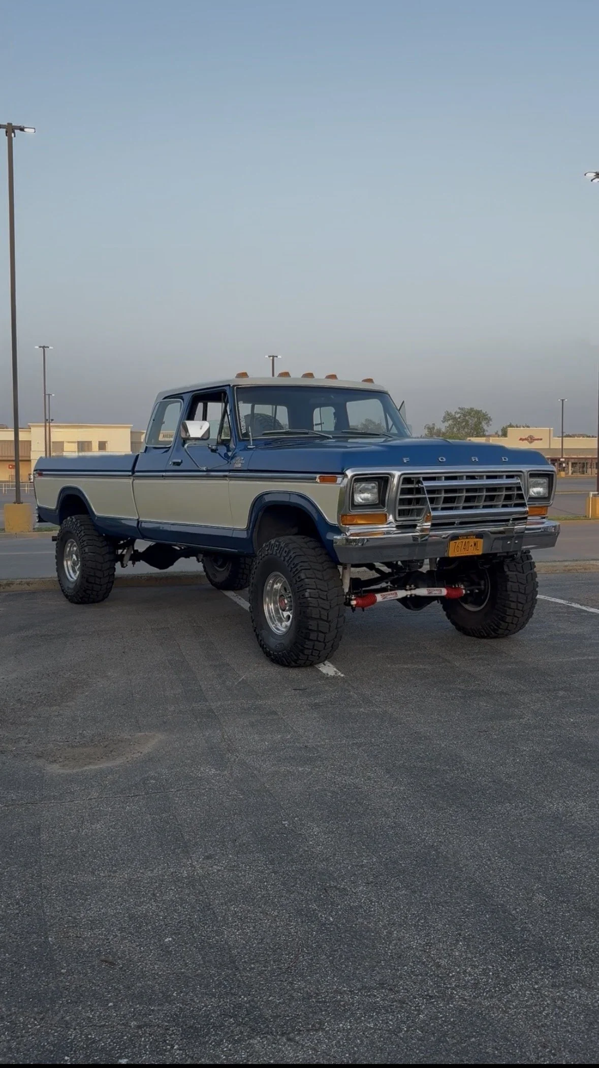 A vintage Ford pickup truck parked in a parking lot, with a two-tone blue and white paint job, large off-road tires, and a raised suspension.