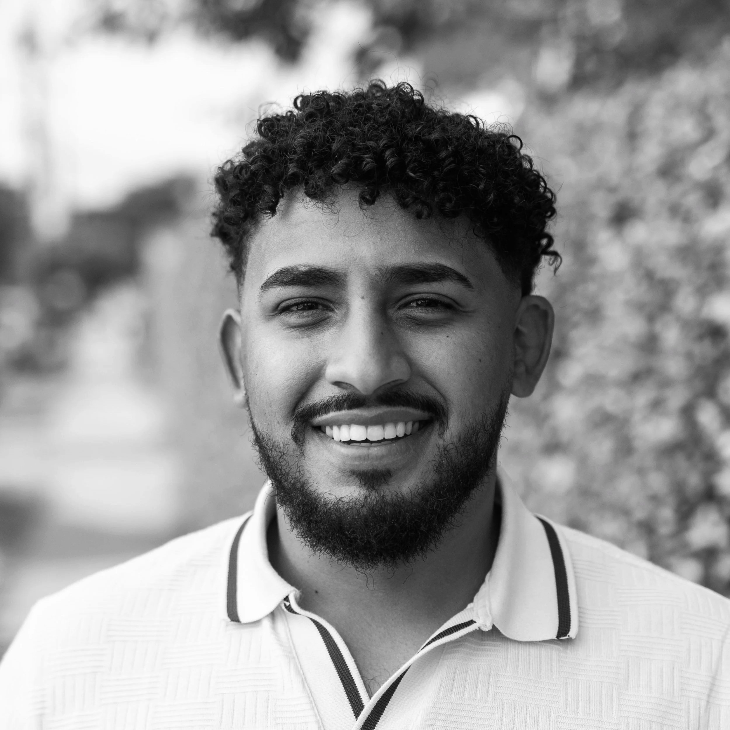 A young man with curly hair and a beard smiling outdoors, wearing a collared shirt.
