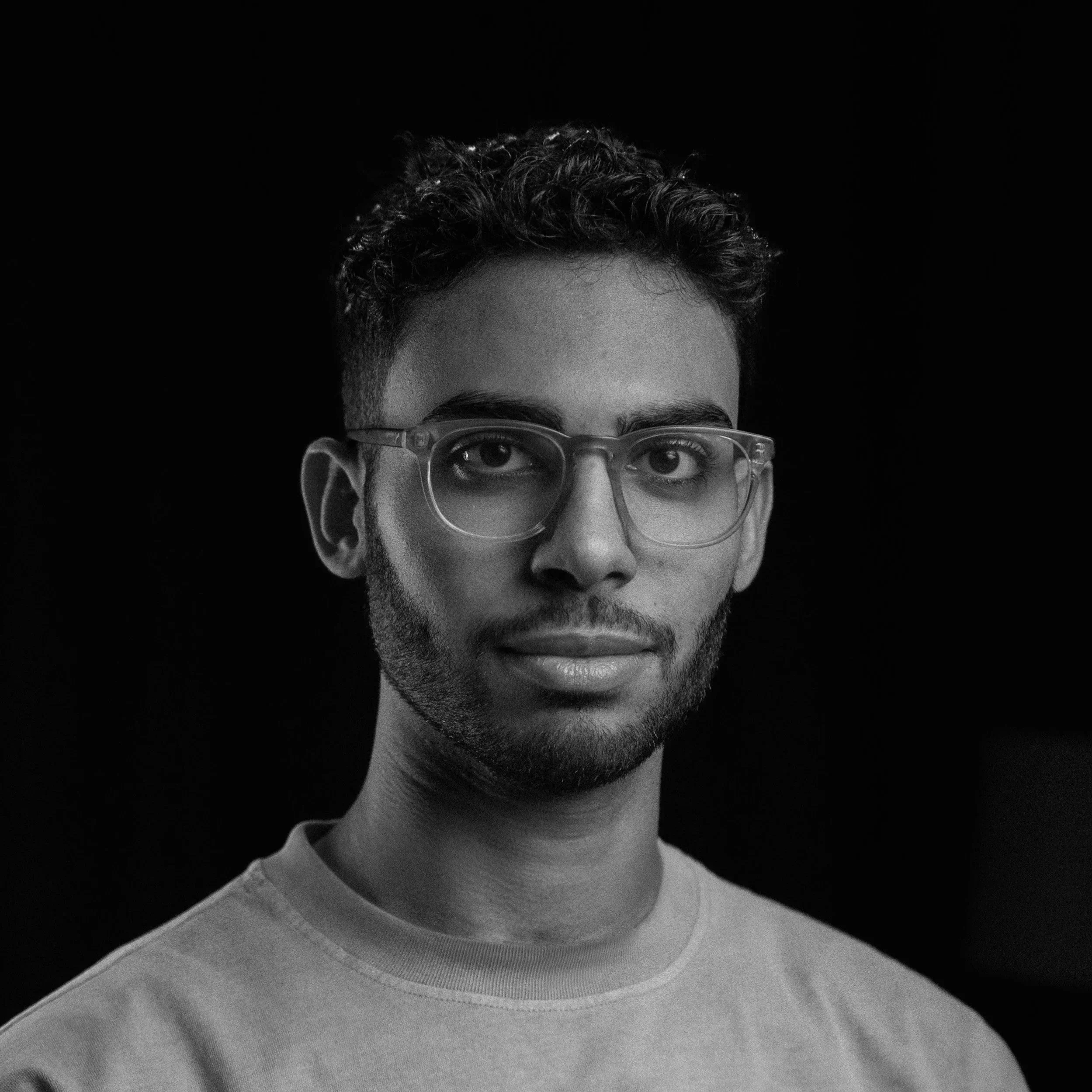 Black and white portrait of a young man with glasses and a beard, wearing a t-shirt, looking directly at the camera.
