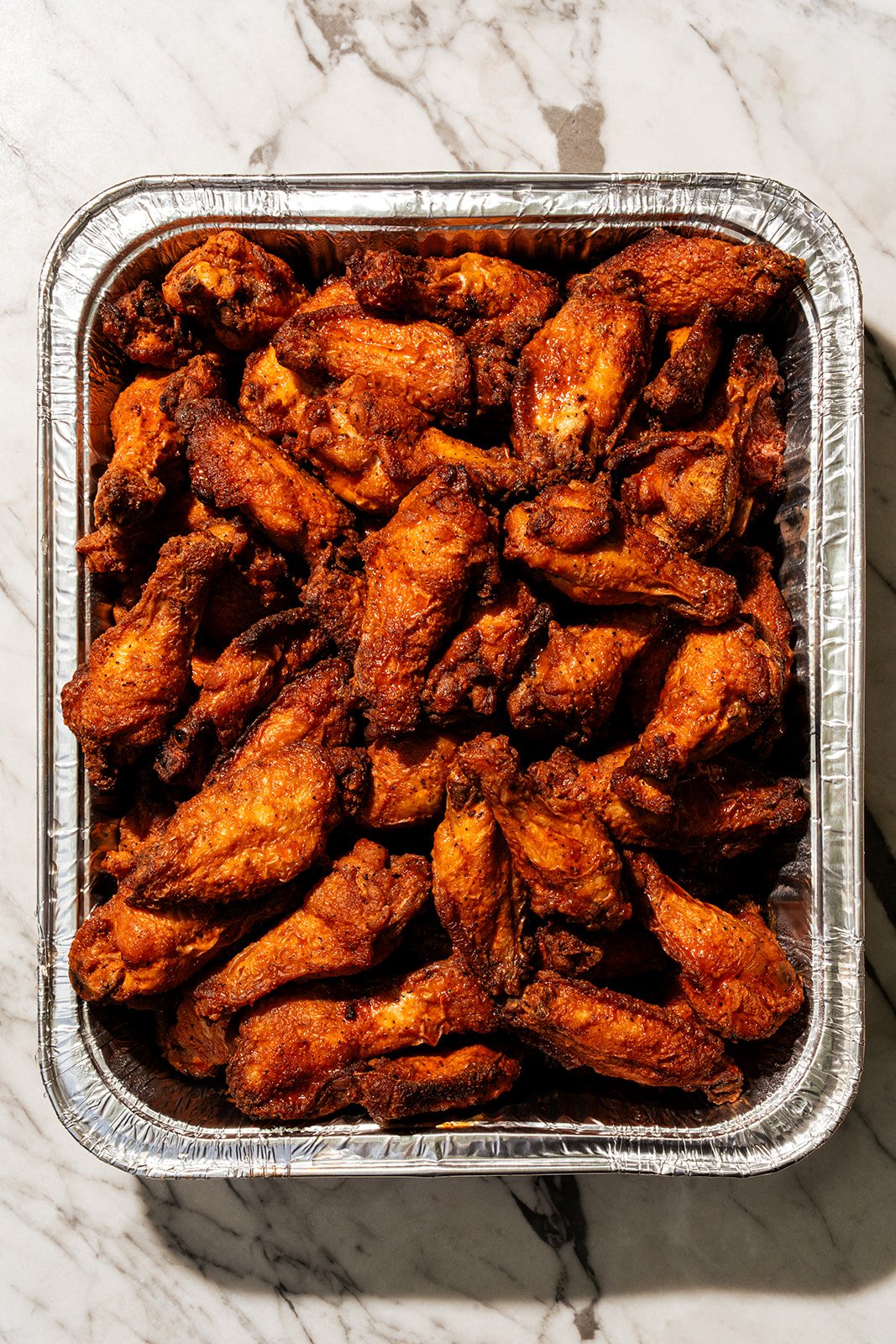 A tray of cooked chicken wings with a crispy, reddish-brown exterior, placed on a marble surface.