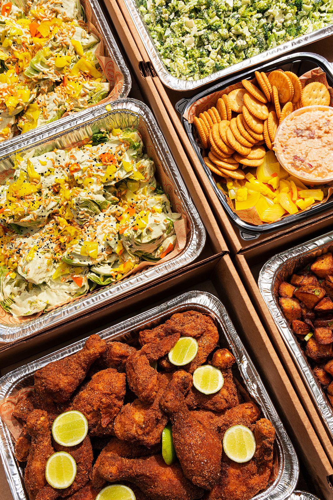 A variety of food trays containing fried chicken, salad, broccoli florets, cheese crackers, and roasted potatoes.