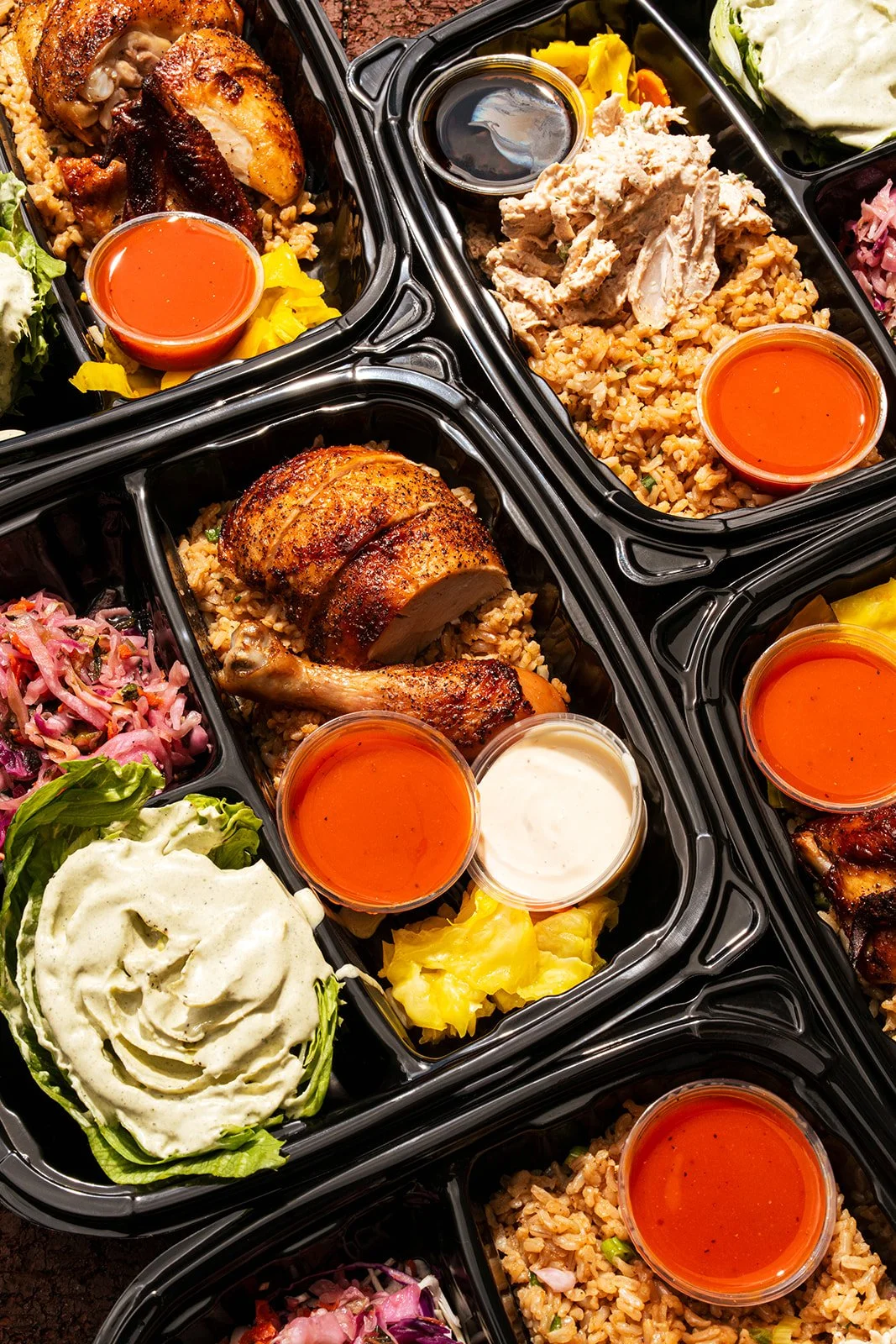 Meal prep containers with roasted chicken, rice, salad, yellow vegetables, and various sauces.