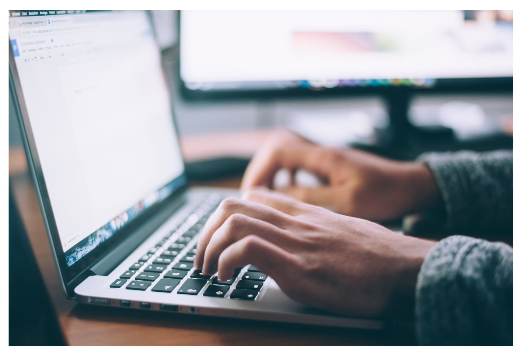 Close-up of hands typing on a laptop keyboard with a monitor in the background.