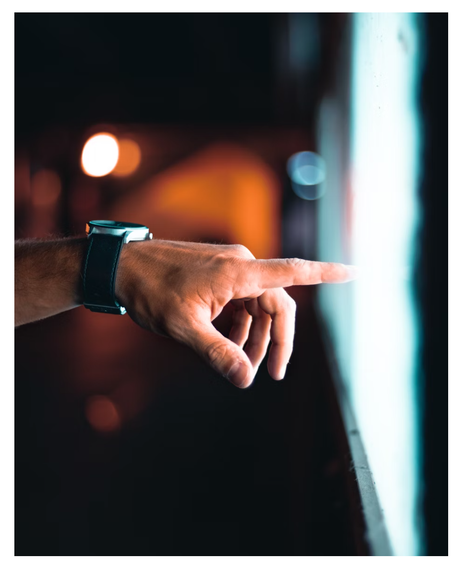 Close-up of a person's hand with a smartwatch, pointing at a screen in a dimly lit environment with blurred lights in the background.
