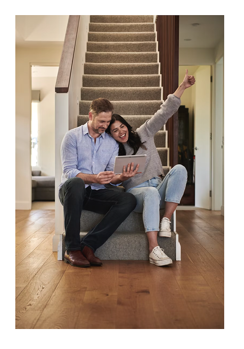A happy couple sits on a staircase landing inside a house, looking at a tablet together. The woman is raising her arm and giving a thumbs-up, while the man is smiling and holding the tablet.