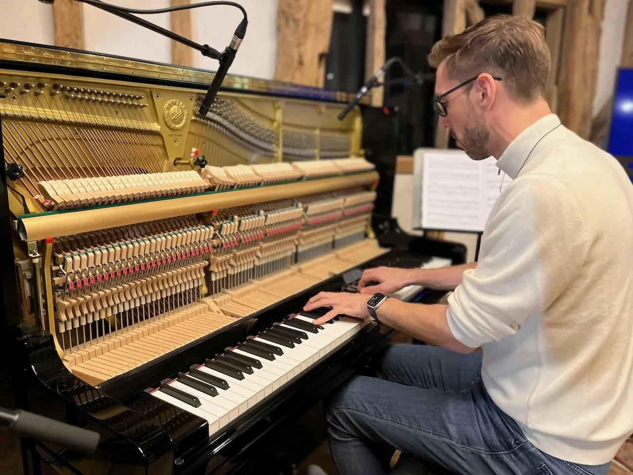 A man wearing glasses and a cream-colored sweater playing an upright piano with its internal strings and hammers visible, in a room with wooden walls and music sheets on a stand.