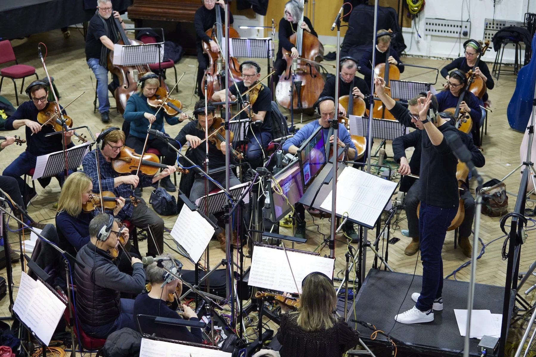 Un orchestre de plusieurs musiciens jouant des instruments à cordes, dirigé par un chef d'orchestre en position centrale, dans une salle de studio d'enregistrement.