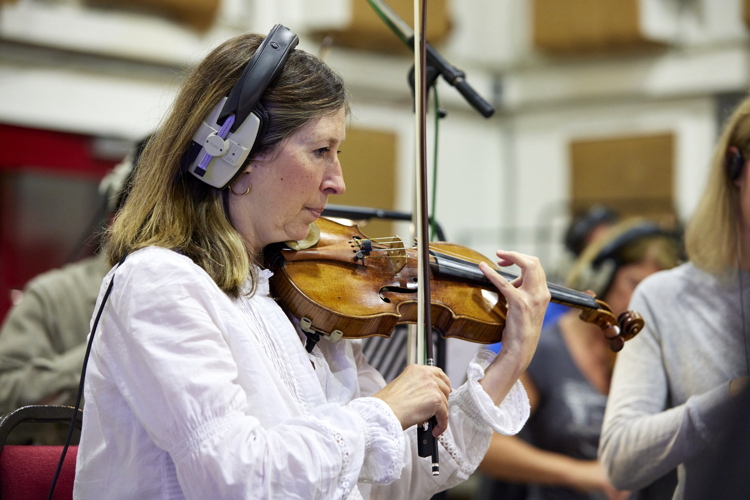 Une femme joue du violon en studio d'enregistrement, portant un casque audio. Autres personnes jouent également d'instruments en arrière-plan.
