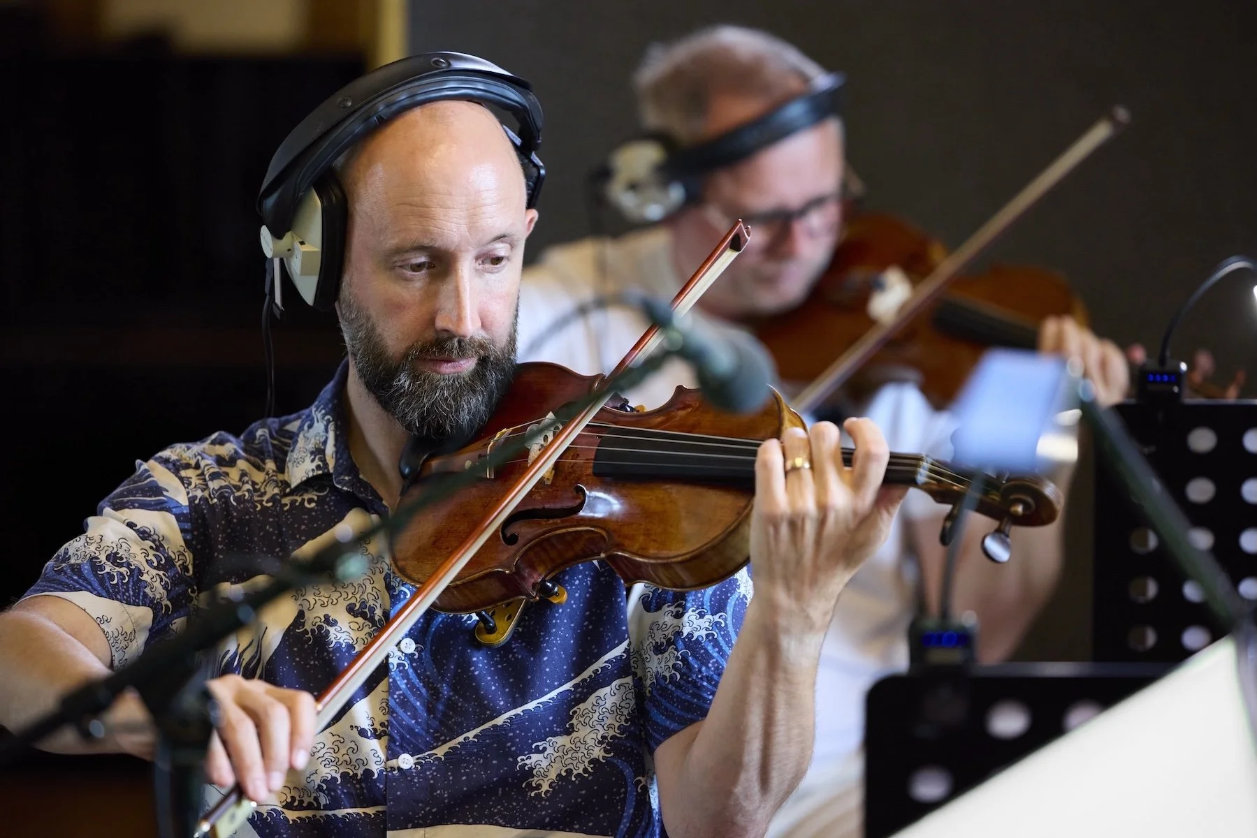 Deux violonistes en studio d'enregistrement, l'un portant un casque audio. L'homme au premier plan joue du violon, concentré, avec un micro devant lui.